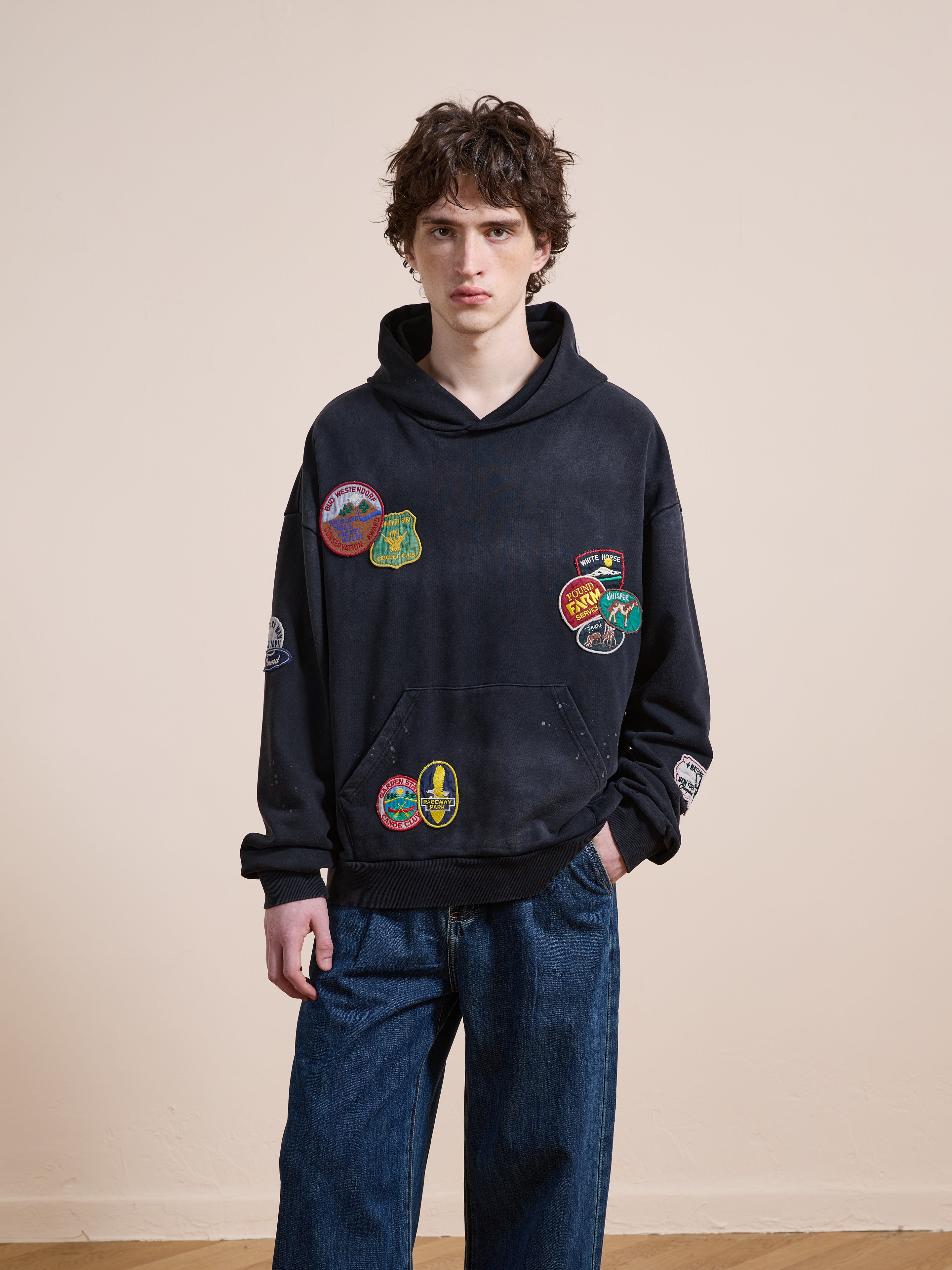 A person wears the FOUND Distressed Patch Hoodie, a vintage washed black hoodie with colorful embroidered patches, paired with loose blue jeans, standing against a plain beige wall.