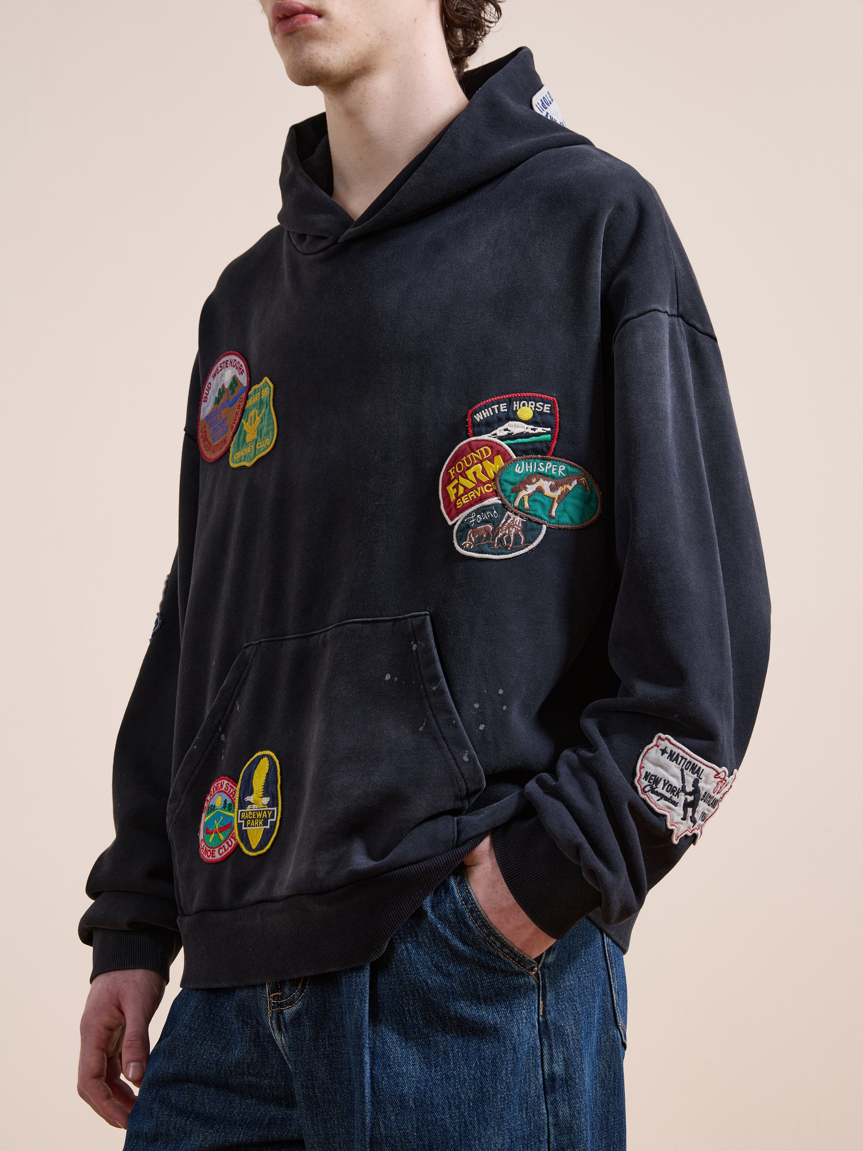 A person wears the FOUND Distressed Patch Hoodie, a vintage washed black unisex relaxed fit top with colorful patches, paired with blue jeans, standing against a plain background.