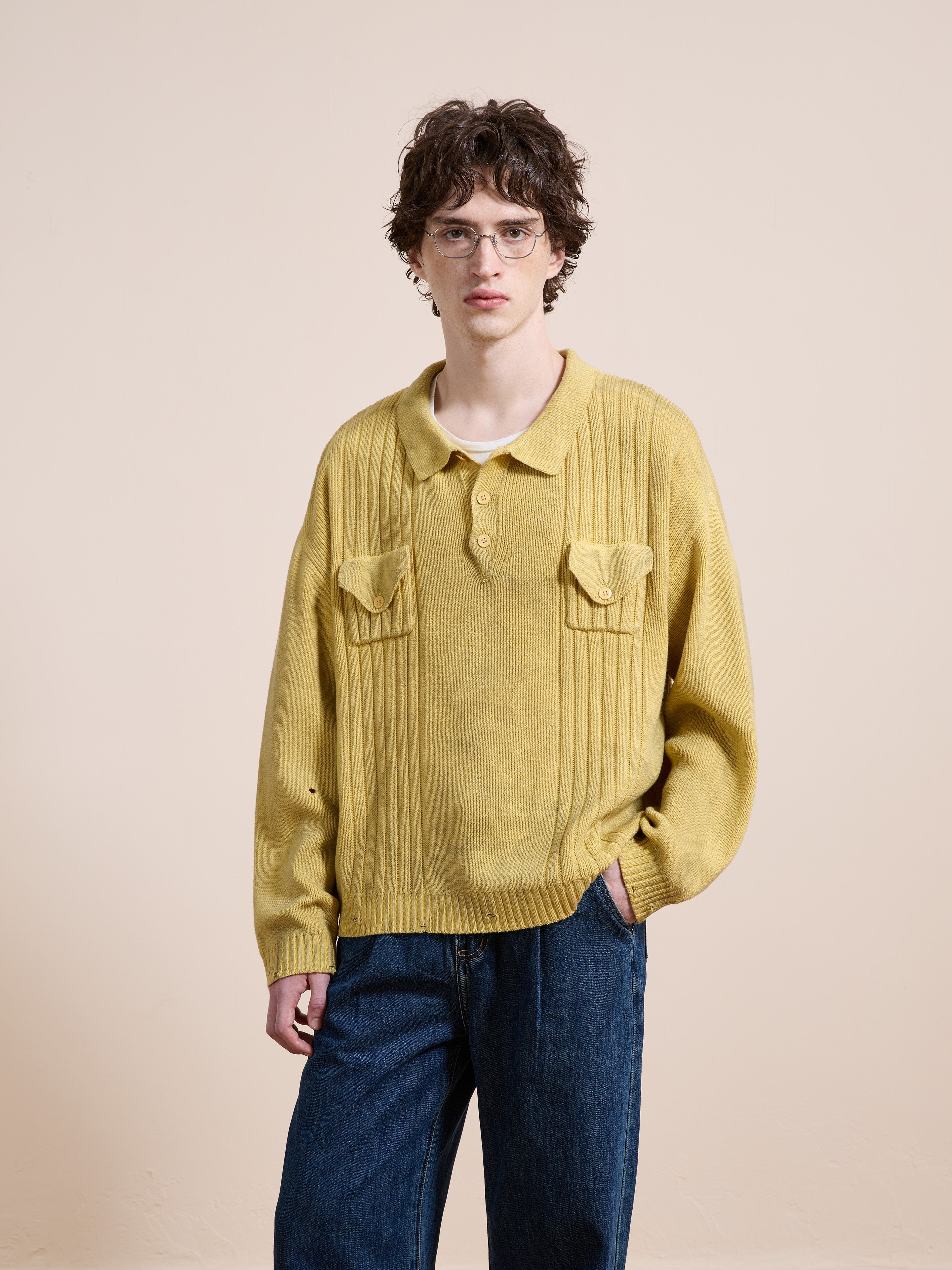 A person with short curly hair and glasses wears the FOUND Distressed Pullover Knit, a yellow unisex sweater with chest pockets, and blue jeans while standing against a plain beige background.