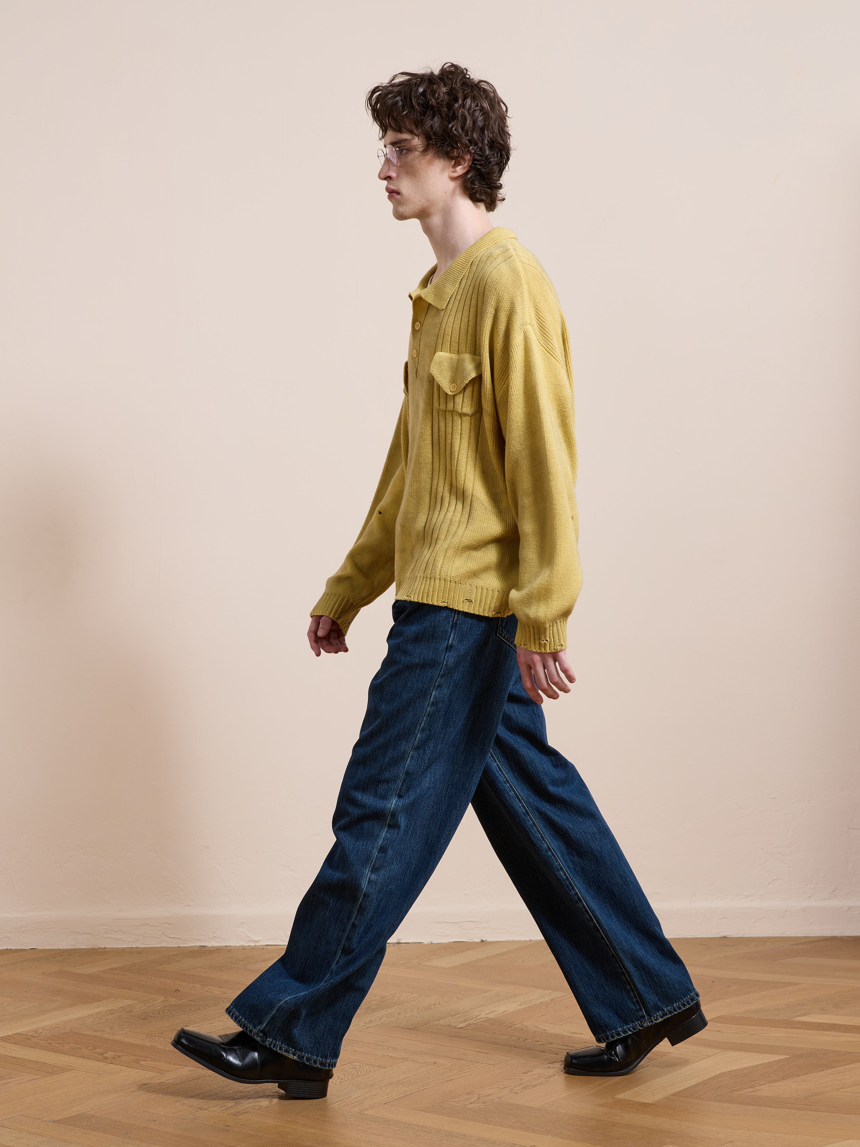 A person wearing the FOUND Distressed Pullover Knit sweater, paired with wide-leg blue jeans and black shoes, walks indoors on a wooden floor against a plain beige wall.