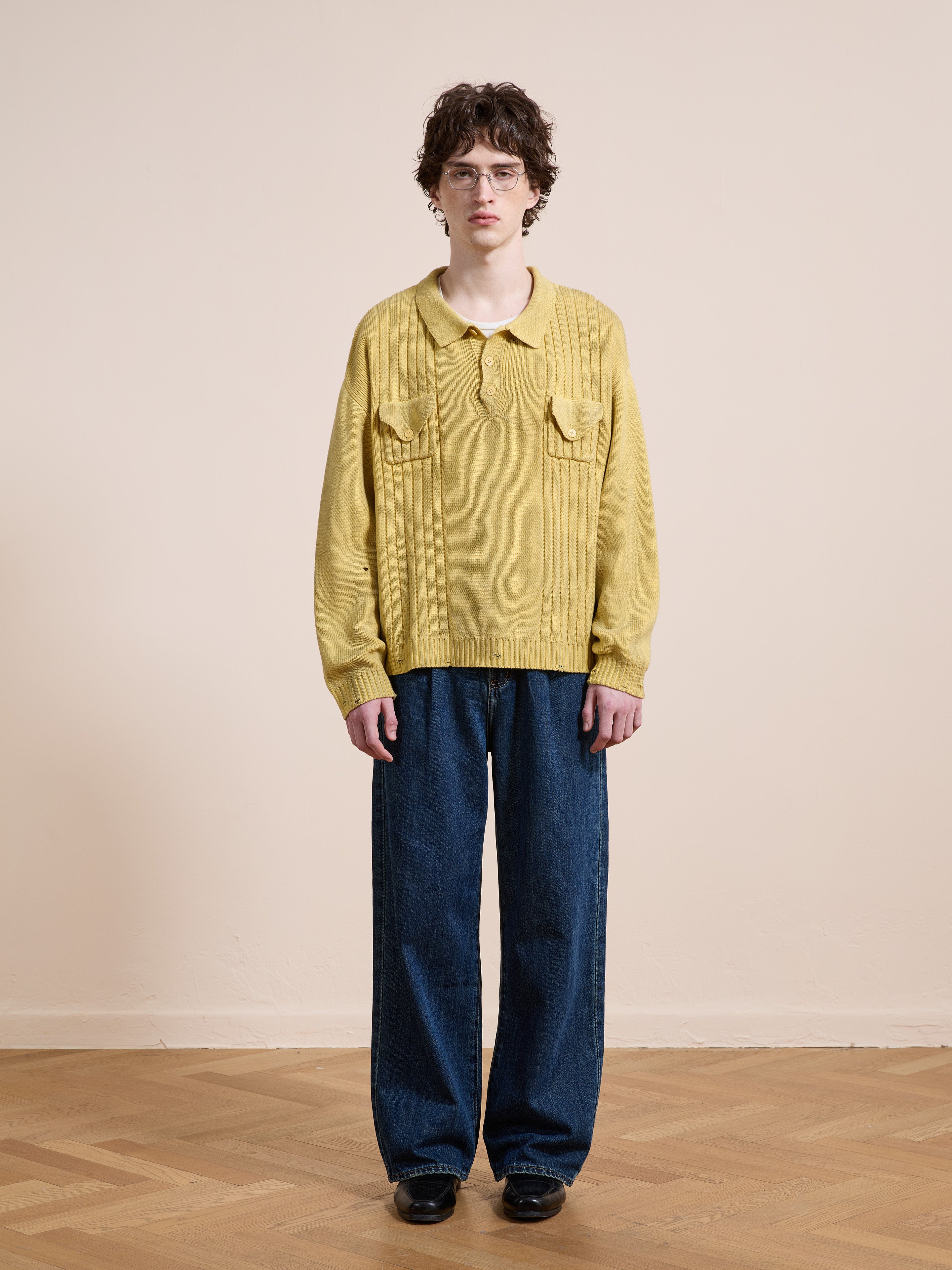 A person stands indoors on a wooden floor against a beige wall, wearing FOUND’s Distressed Pullover Knit sweater in yellow, paired with blue wide-leg jeans and black shoes, showcasing versatile transitional knitwear for any season.