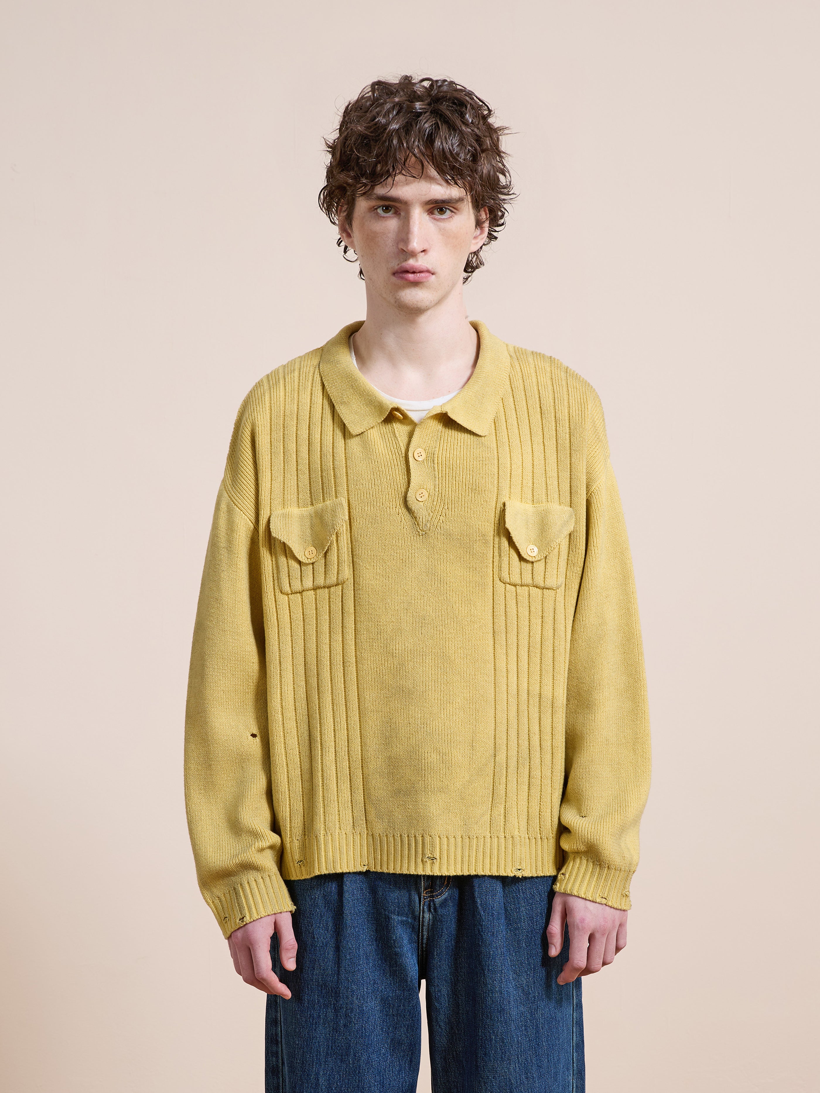 A person with curly brown hair wears the FOUND Distressed Pullover Knit in yellow and blue jeans, standing against a plain beige background.