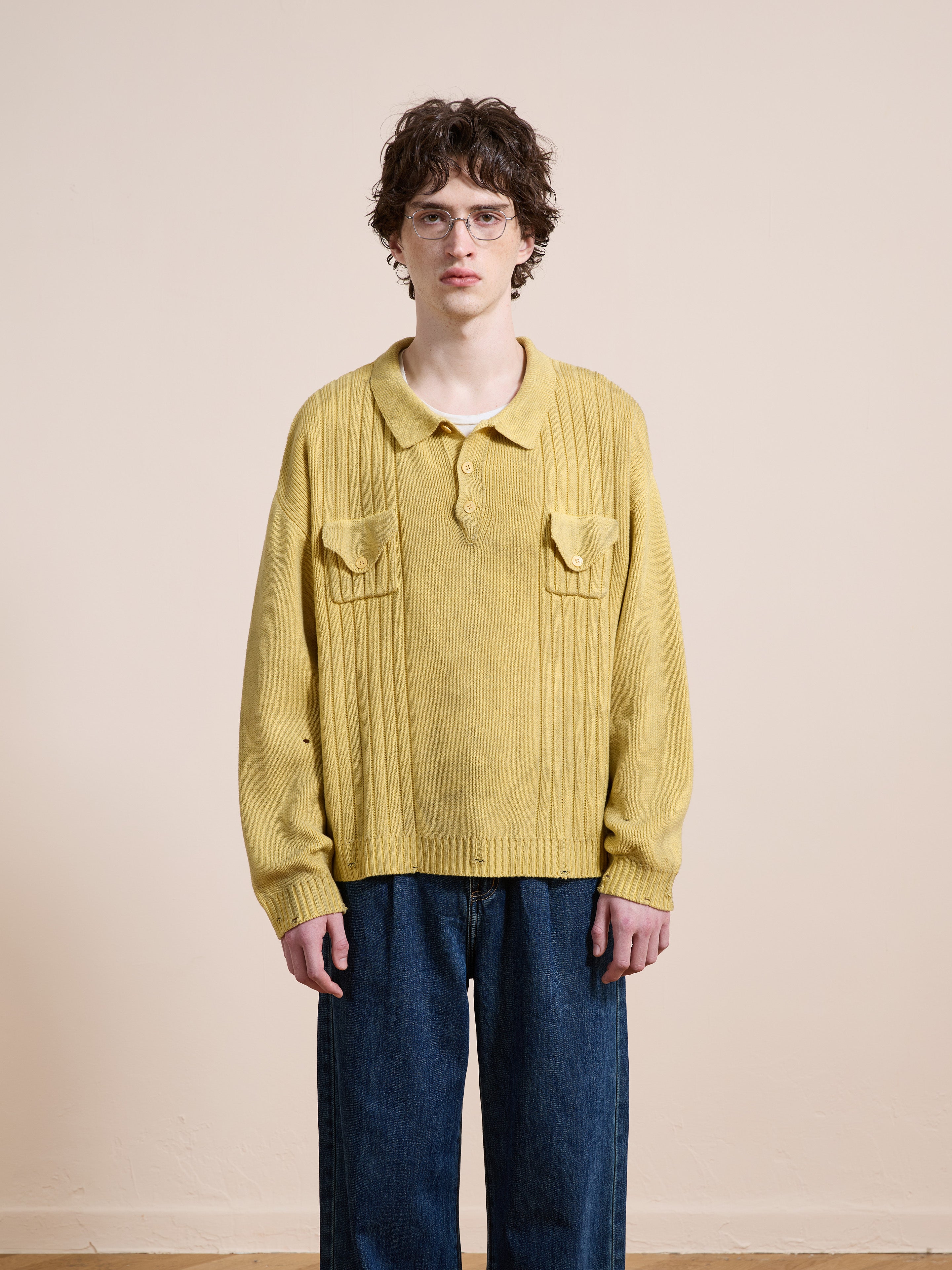 A person with short curly hair and glasses wears the FOUND Distressed Pullover Knit in yellow and loose blue jeans, standing against a plain beige background.