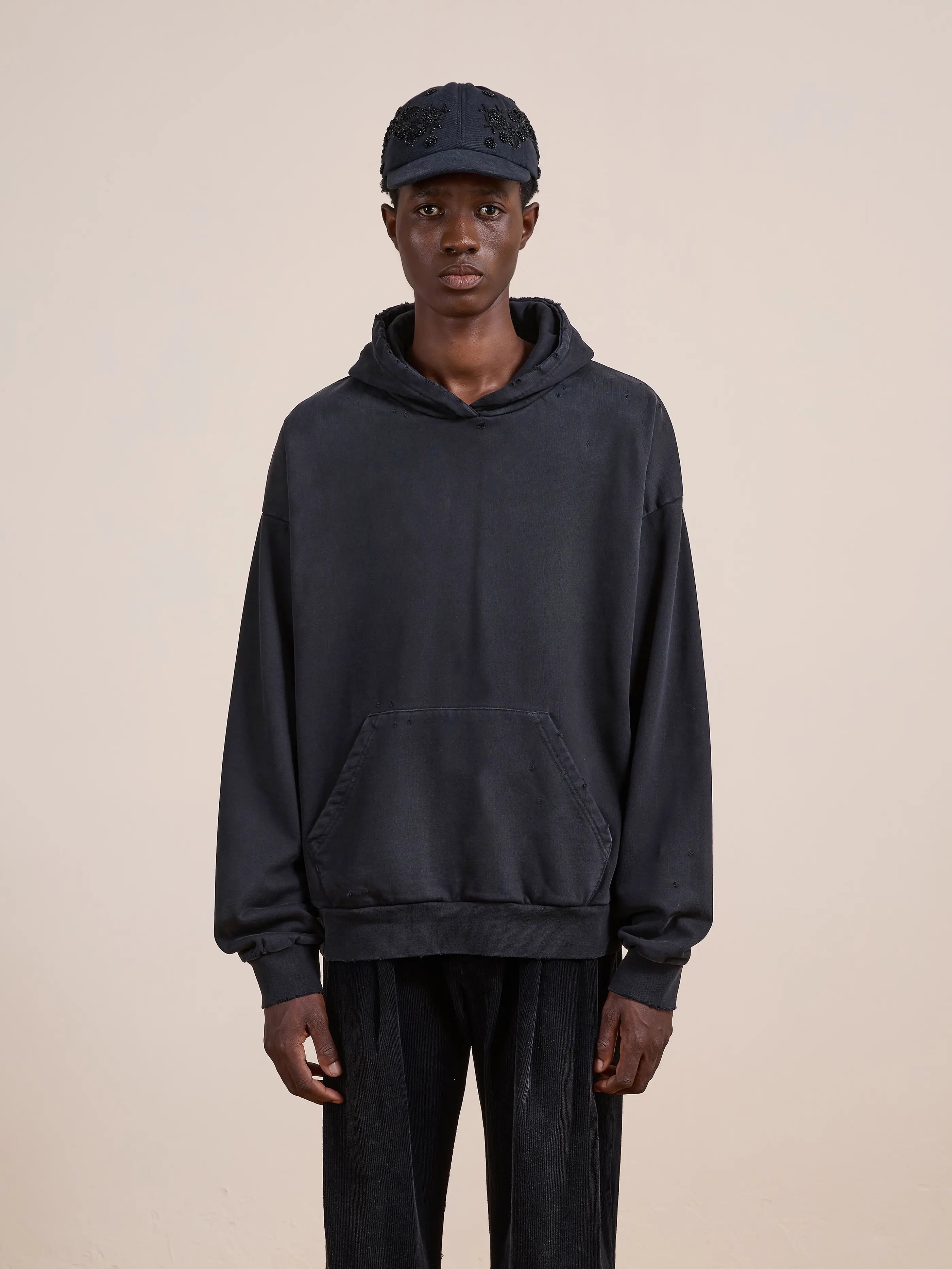 A person faces forward in a FOUND Distressed Pullover Hoodie with a unisex relaxed fit, paired with black pants and a black cap, set against a plain light background.