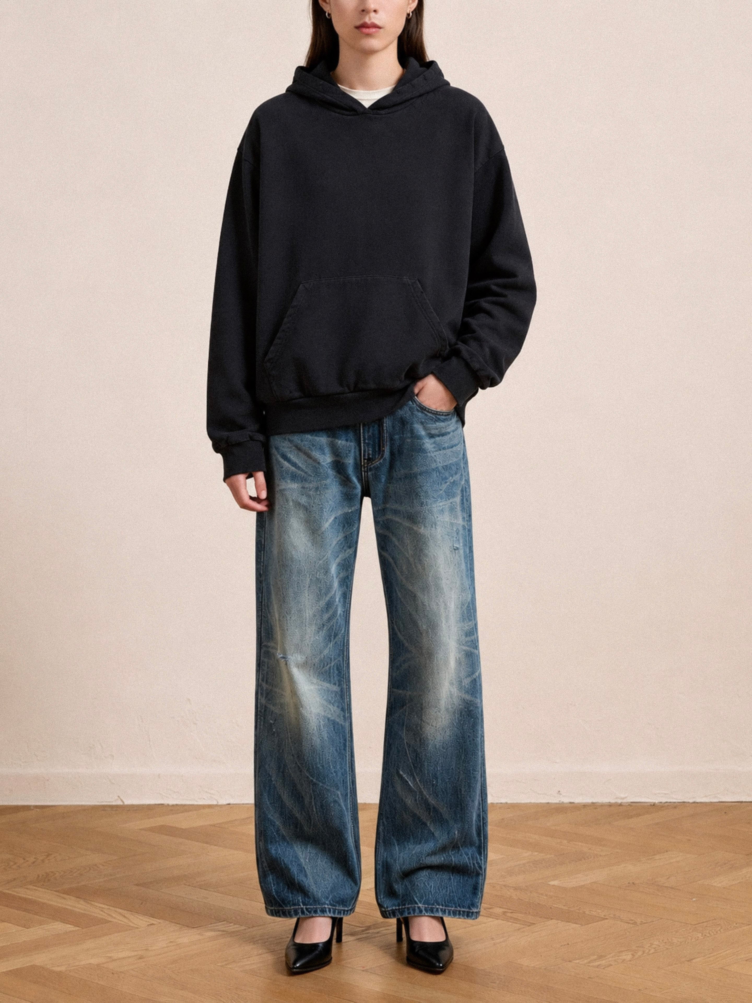 A person stands indoors wearing the FOUND Distressed Pullover Hoodie with a unisex relaxed fit, paired with loose blue jeans and black heels on a wooden floor against a plain light-colored wall.