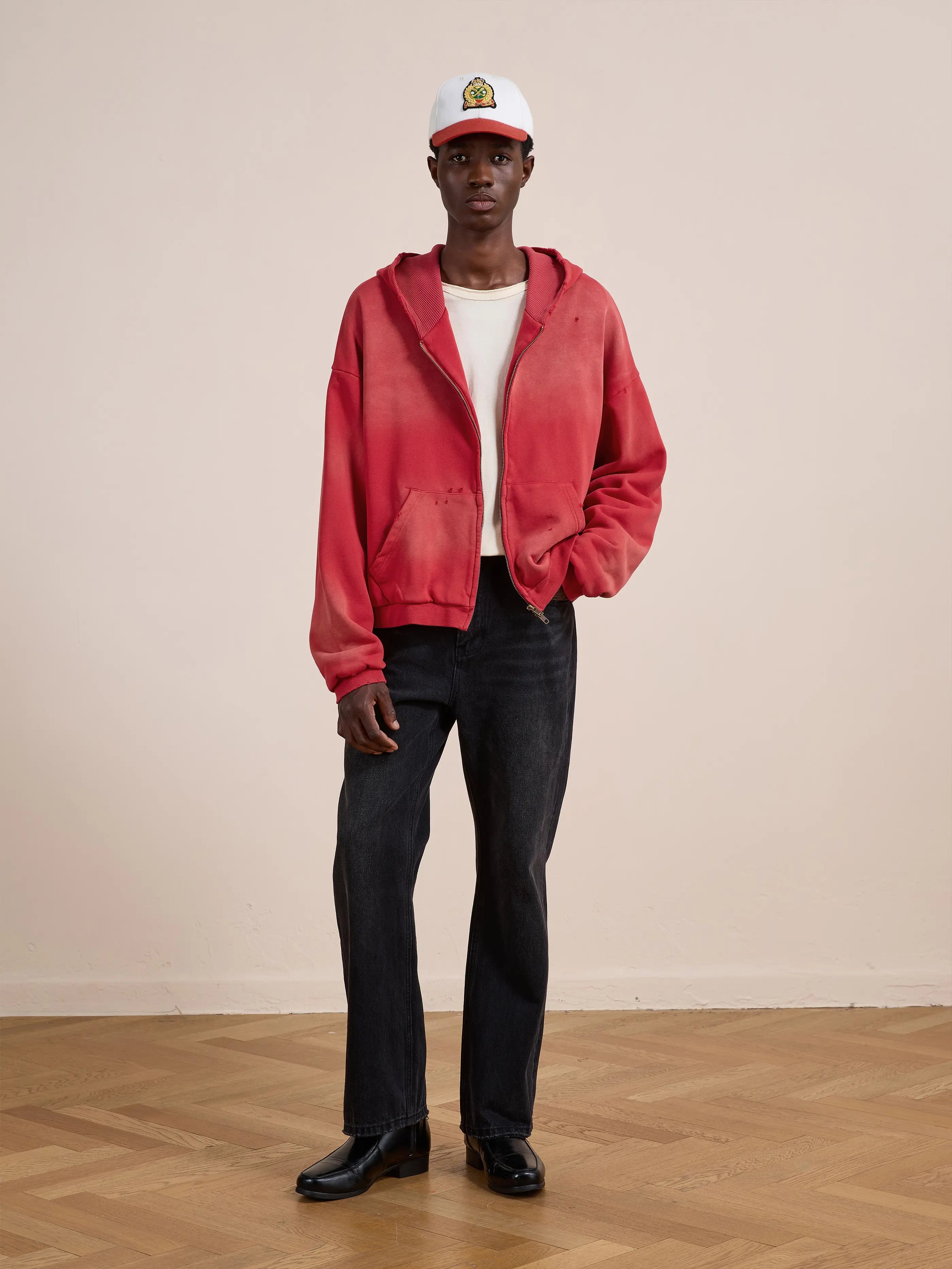 Indoors, a person wears a white cap, FOUND Distressed Thermal Lined Zip Up Hoodie in red, white t-shirt, black jeans, and black boots on a wooden floor against a plain light wall.