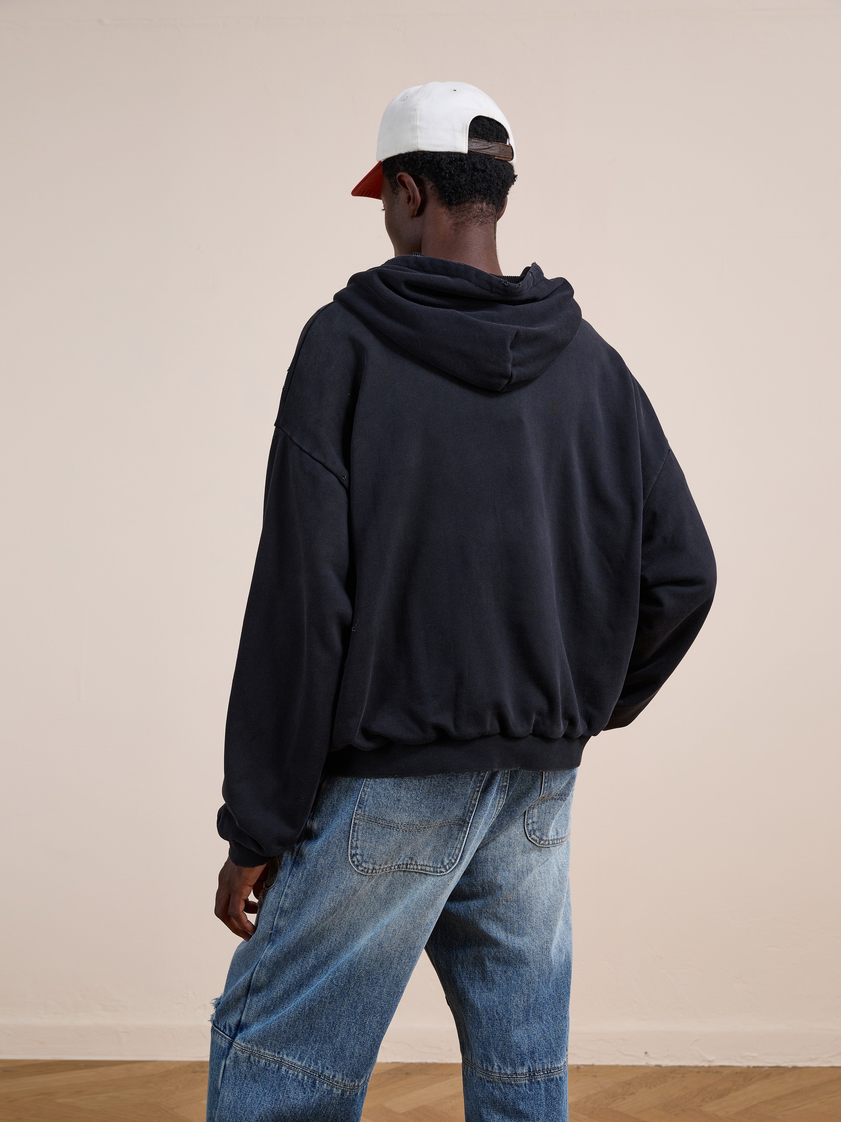 Someone wearing the FOUND Distressed Thermal Lined Zip Up Hoodie in black, blue jeans, and a two-tone baseball cap stands facing away from the camera in a neutral indoor setting.