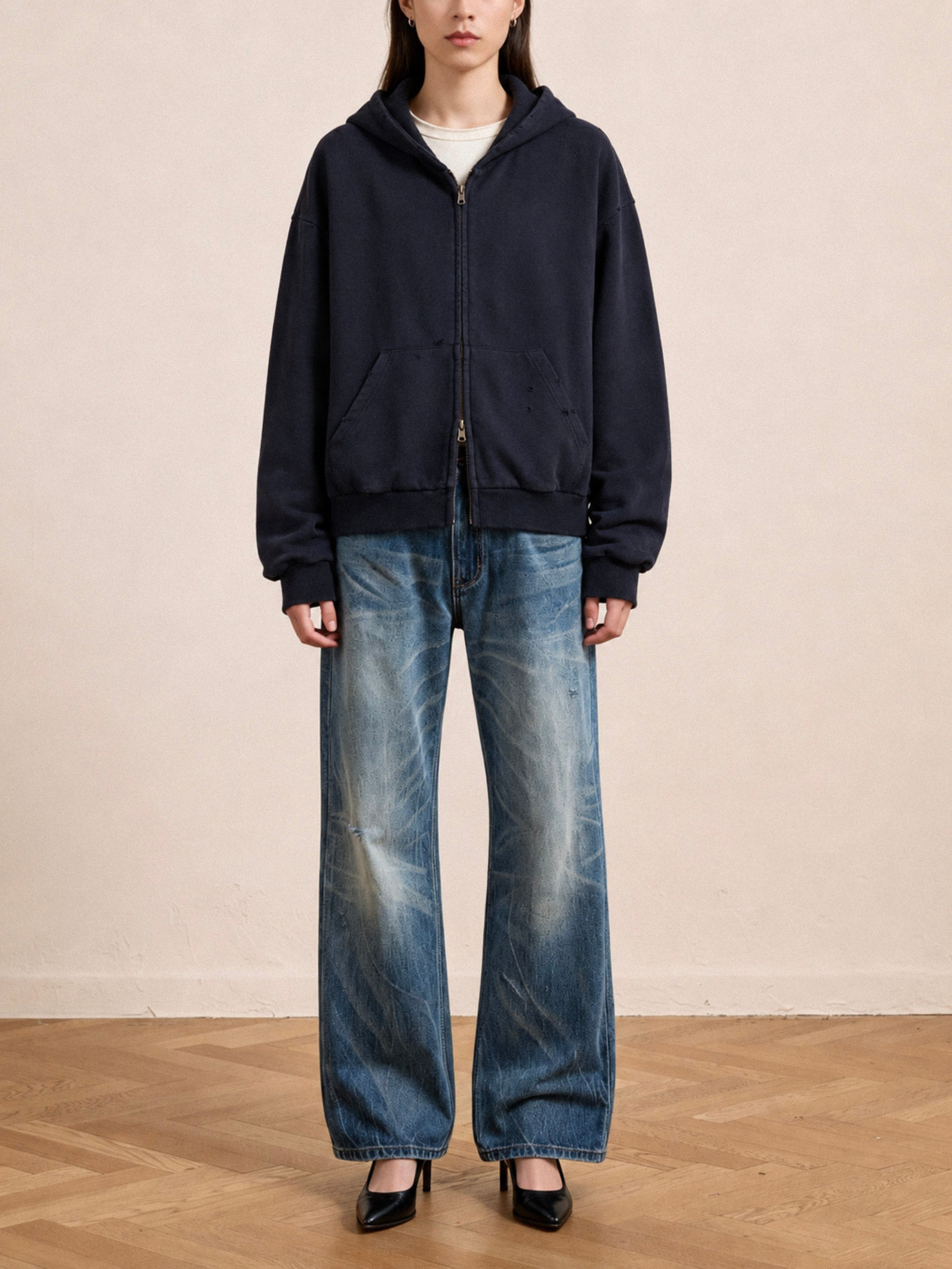 A person stands indoors in a FOUND Distressed Thermal Lined Zip Up Hoodie over a light shirt, wide-leg faded jeans, and black heels, on a wooden floor with a neutral background.