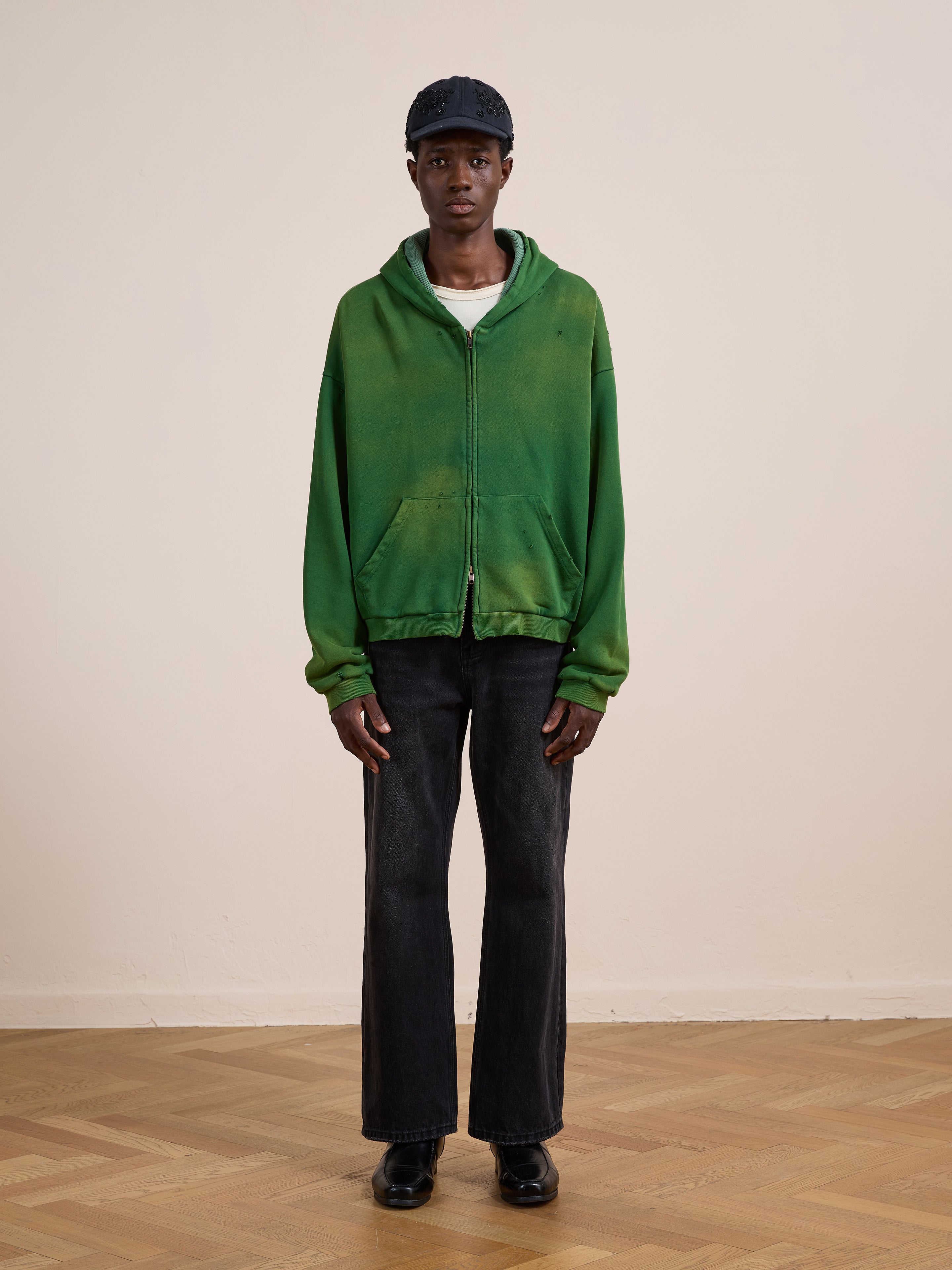 A person stands indoors on a wooden floor, facing the camera against a plain background, wearing a green FOUND Distressed Thermal Lined Zip Up Hoodie, dark jeans, black shoes, a white shirt, and a dark cap.