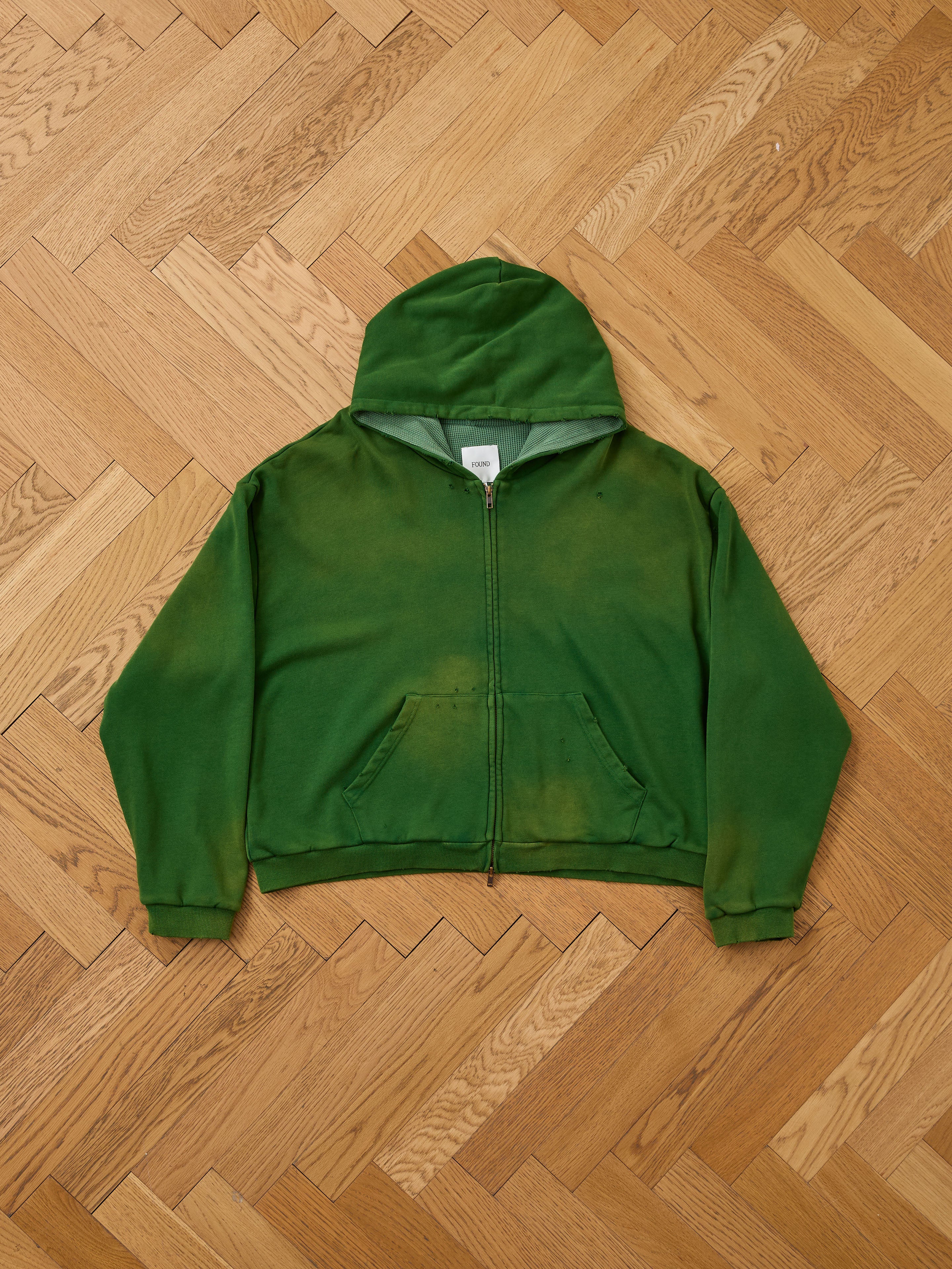 The FOUND Distressed Thermal Lined Zip Up Hoodie, a green unisex relaxed fit with hood and pockets, is laid flat on a herringbone wooden floor.
