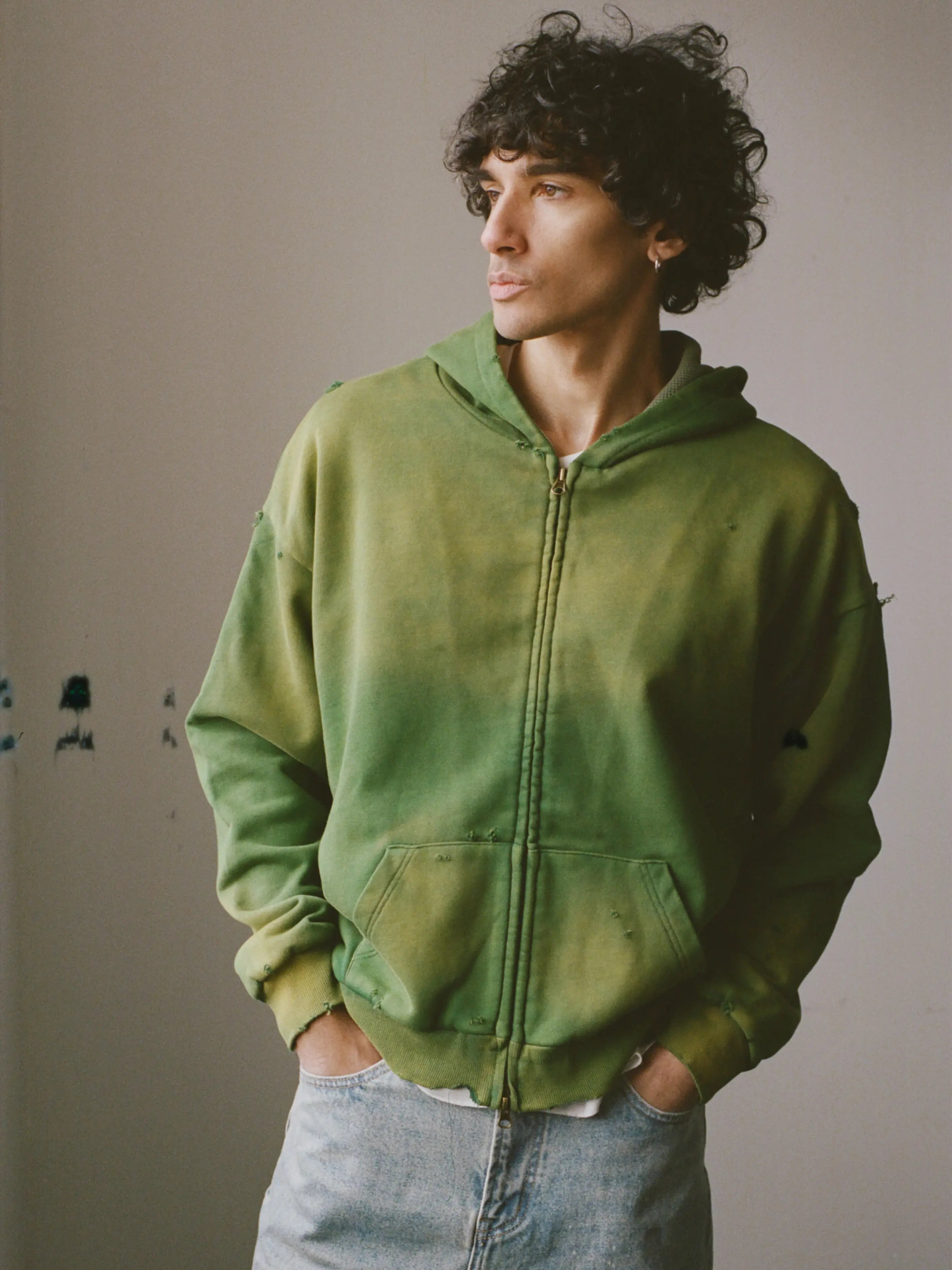 A person with curly hair wears a FOUND Distressed Thermal Lined Zip Up Hoodie in green and light blue jeans, standing against a plain wall with hands in pockets, looking to the side.