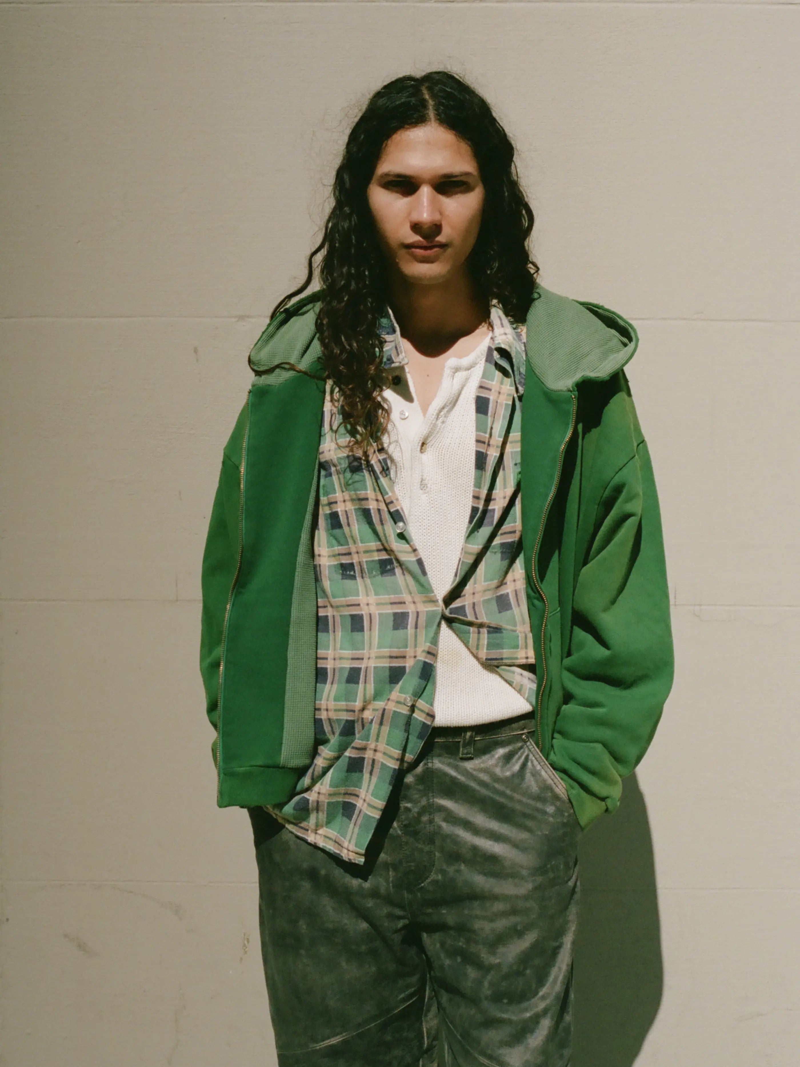 A person with long curly hair stands against a plain wall, wearing a green FOUND Distressed Thermal Lined Zip Up Hoodie over a plaid shirt and white top, paired with grey pants and has hands casually tucked in pockets.