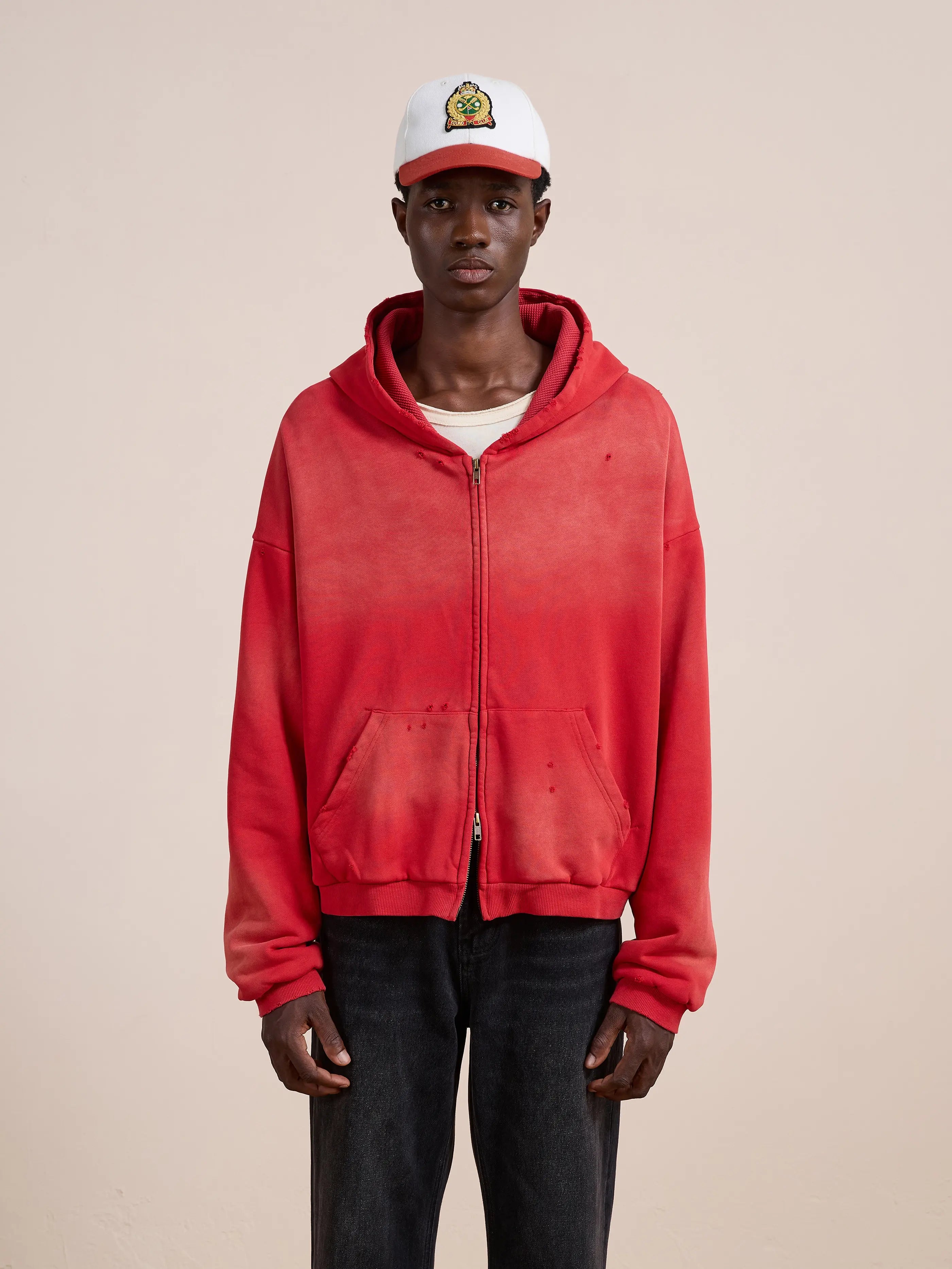 A person wears a red FOUND Distressed Thermal Lined Zip Up Hoodie, white T-shirt, black jeans, and a white baseball cap while standing against a plain light background.