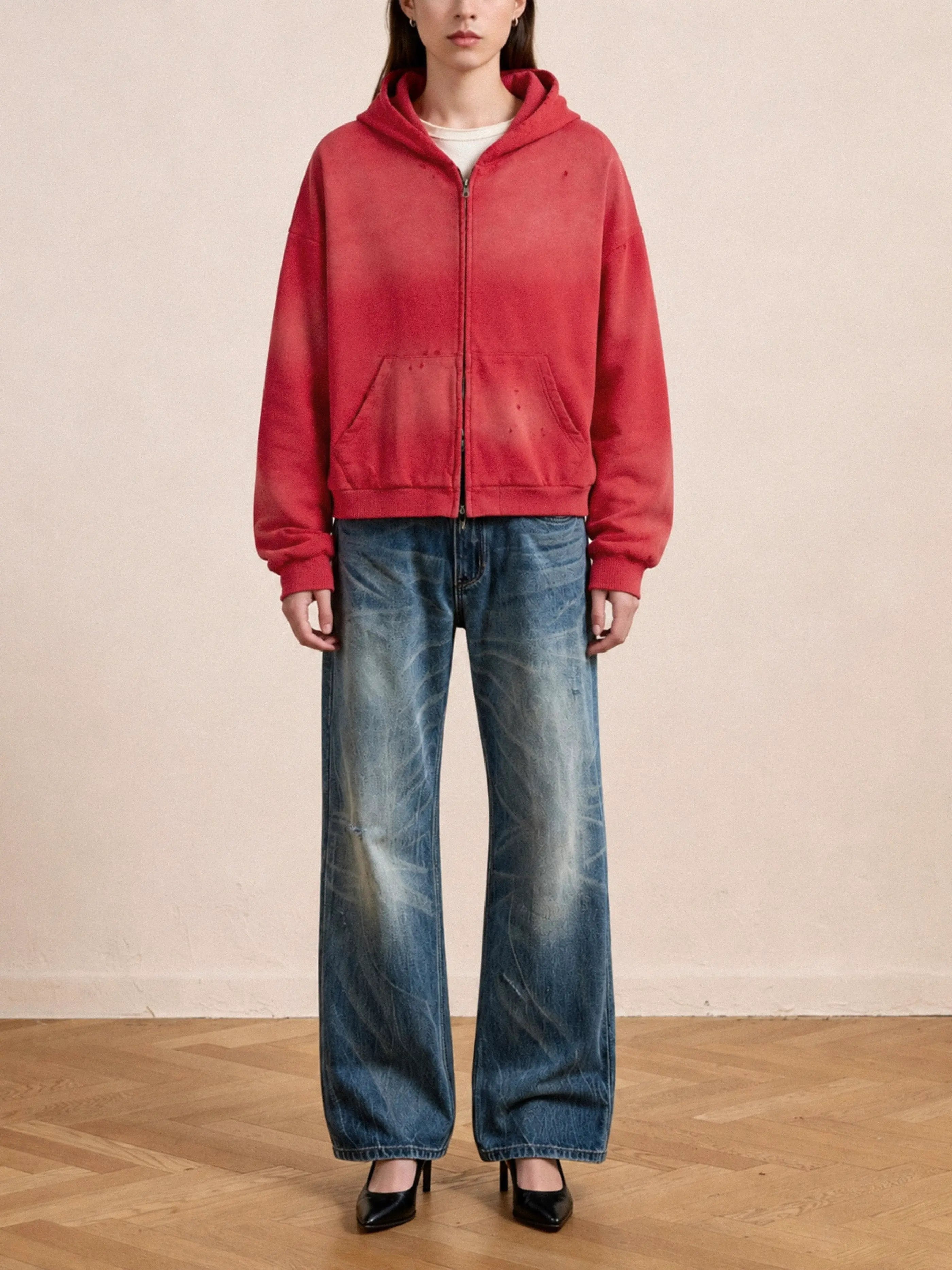 A person stands indoors facing forward against a plain background, wearing the FOUND Distressed Thermal Lined Zip Up Hoodie in faded red, paired with wide-leg distressed blue jeans and black heels.