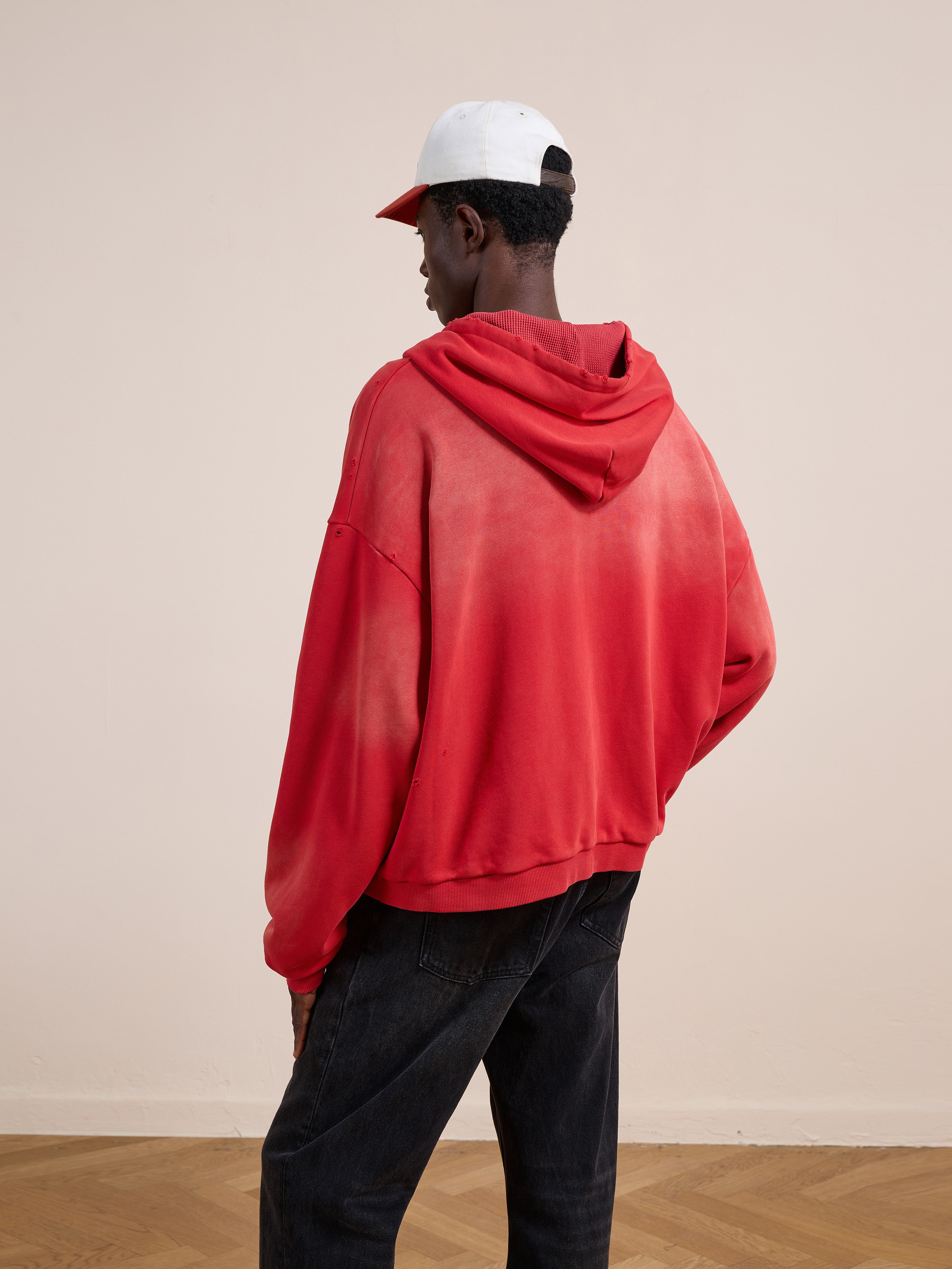 A person wearing a red FOUND Distressed Thermal Lined Zip Up Hoodie, black jeans, and a white and red cap stands facing away from the camera in a neutral indoor setting.