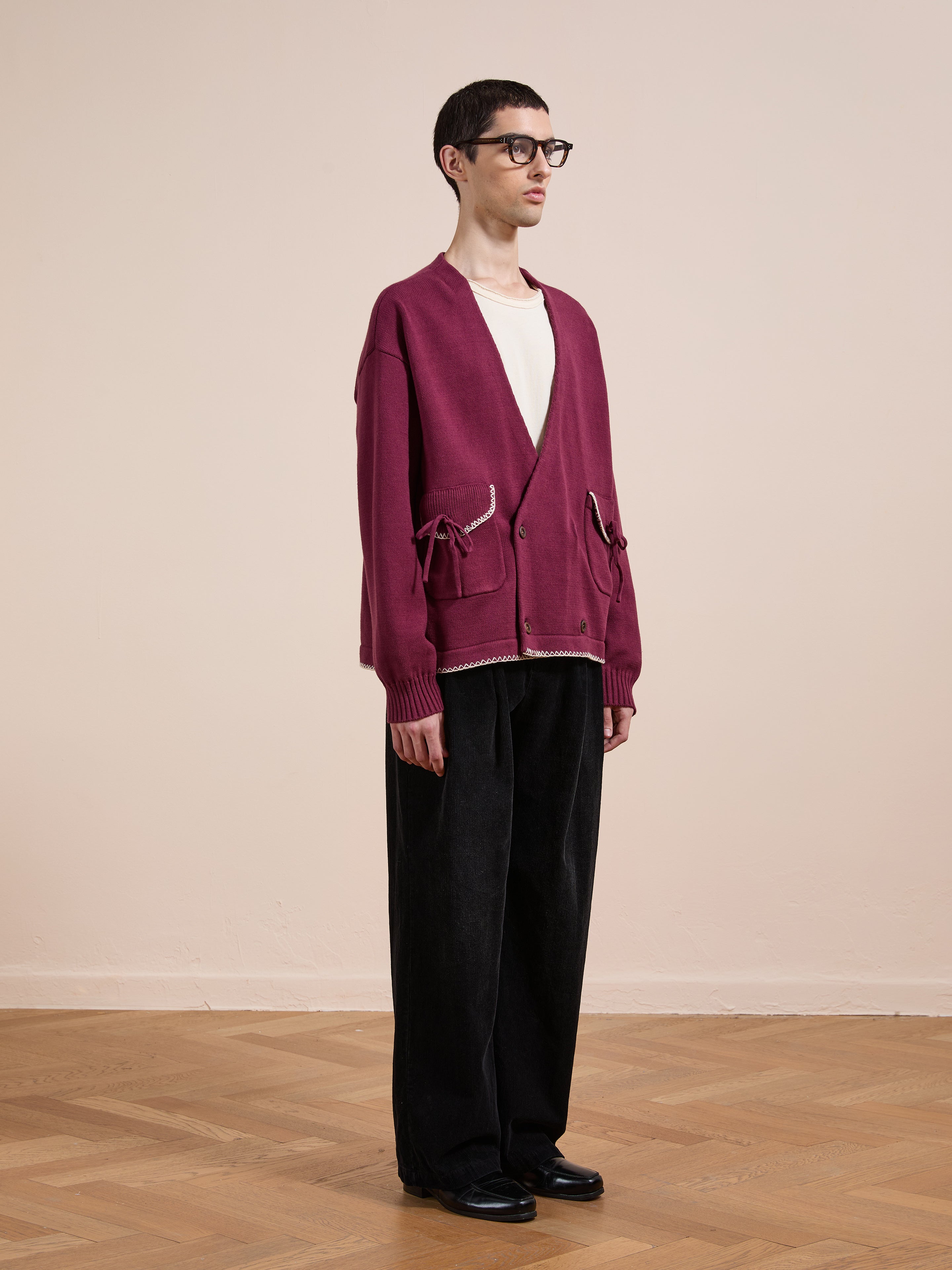 A person stands indoors against a beige wall wearing FOUND's Double Breasted Bow Cardigan in burgundy, paired with a white t-shirt, black wide-leg pants, black shoes, and glasses.
