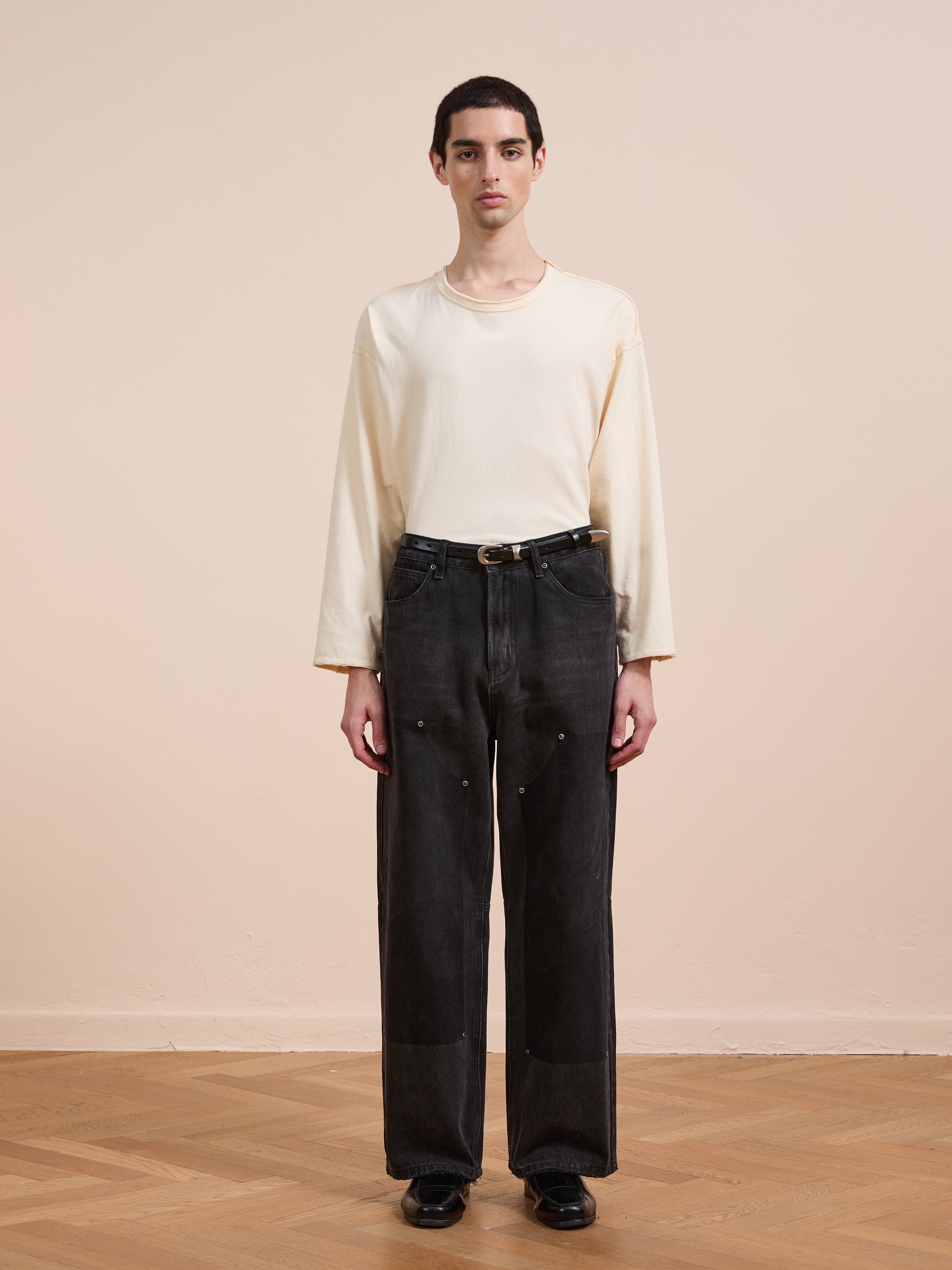 Indoors on a wooden floor, a person wears a loose cream long-sleeve shirt, black FOUND Double Knee Carpenter Jeans with a belt, and black shoes, standing against a plain beige wall.