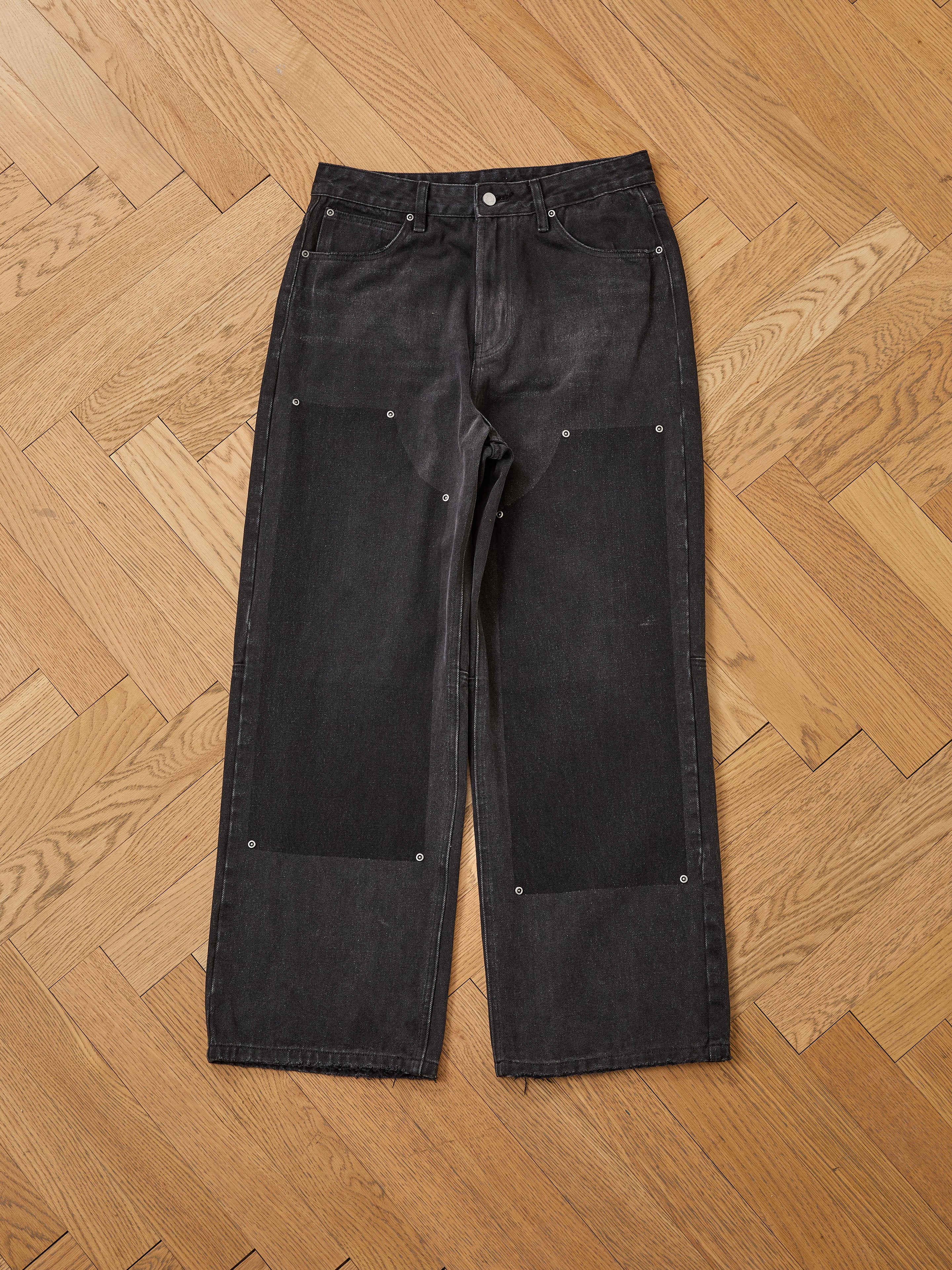 The FOUND Double Knee Carpenter Jeans feature a relaxed fit, wide legs, large black thigh patches, and silver rivets, shown here laid flat on a wooden parquet floor.
