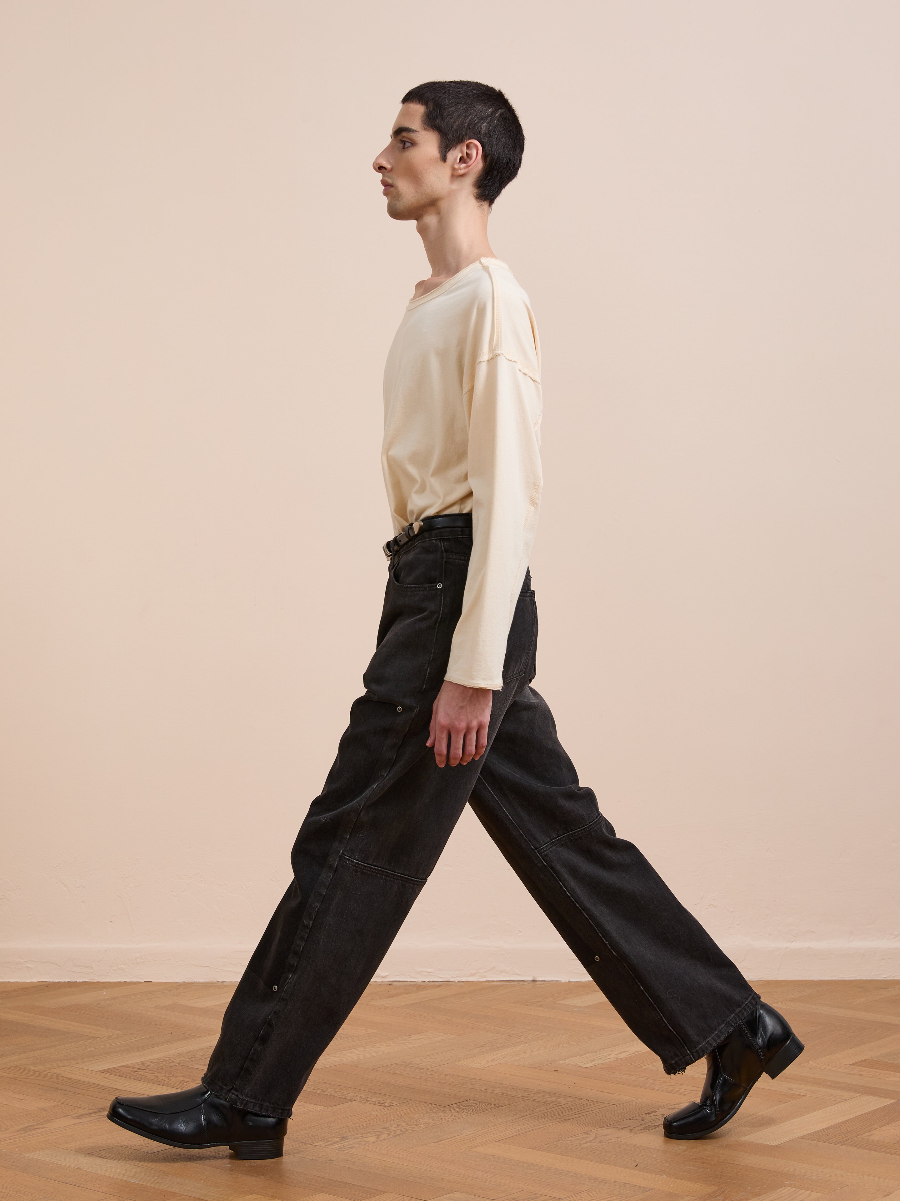 A person with short dark hair wearing a cream long-sleeve shirt, FOUND Double Knee Carpenter Jeans, and black boots walks indoors on a wooden floor against a beige wall.