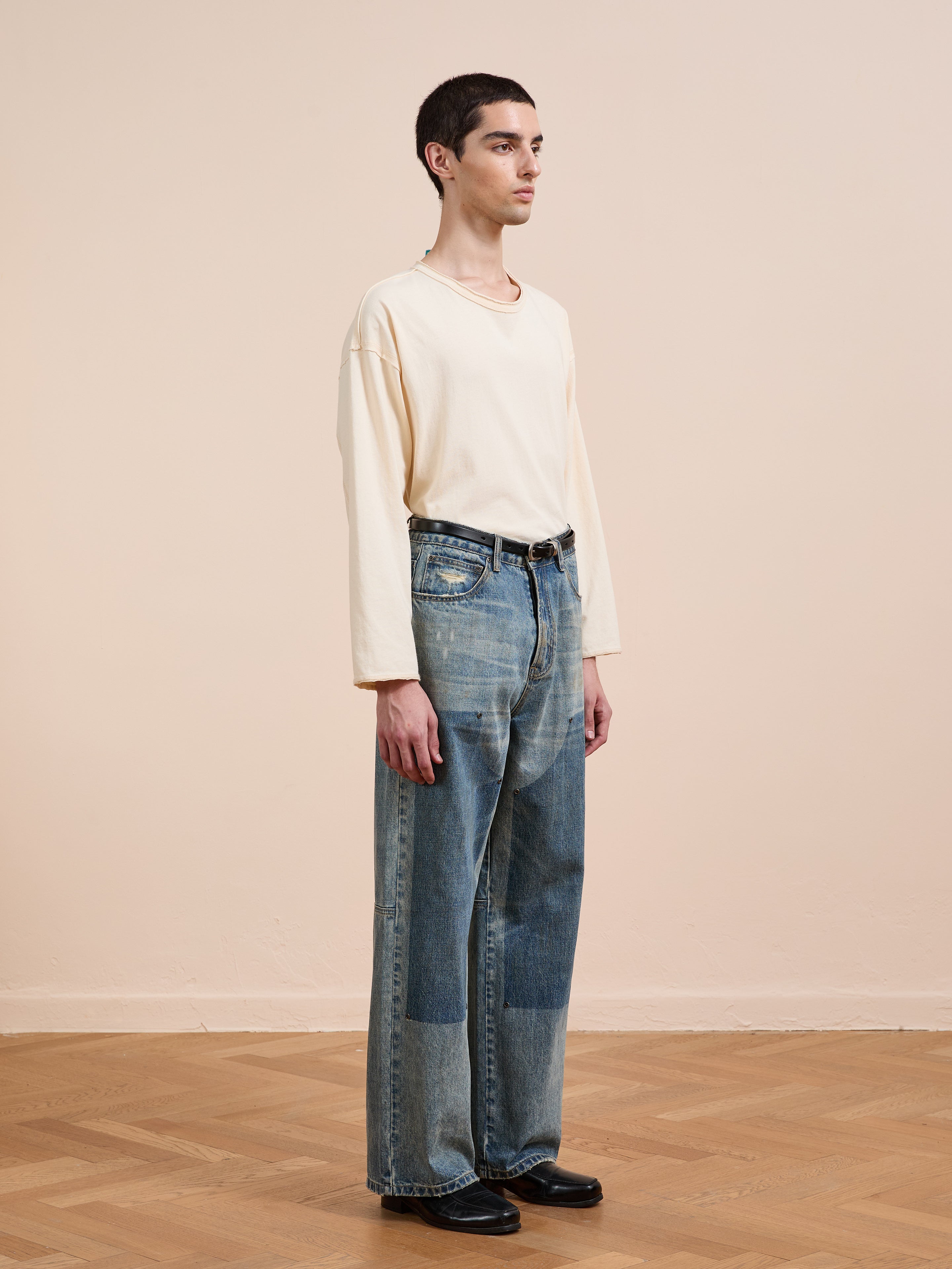 A person stands indoors on a wooden floor, wearing a loose cream long-sleeve shirt, black shoes, and FOUND Double Knee Carpenter Jeans in a relaxed vintage wash, facing right against a plain beige wall.