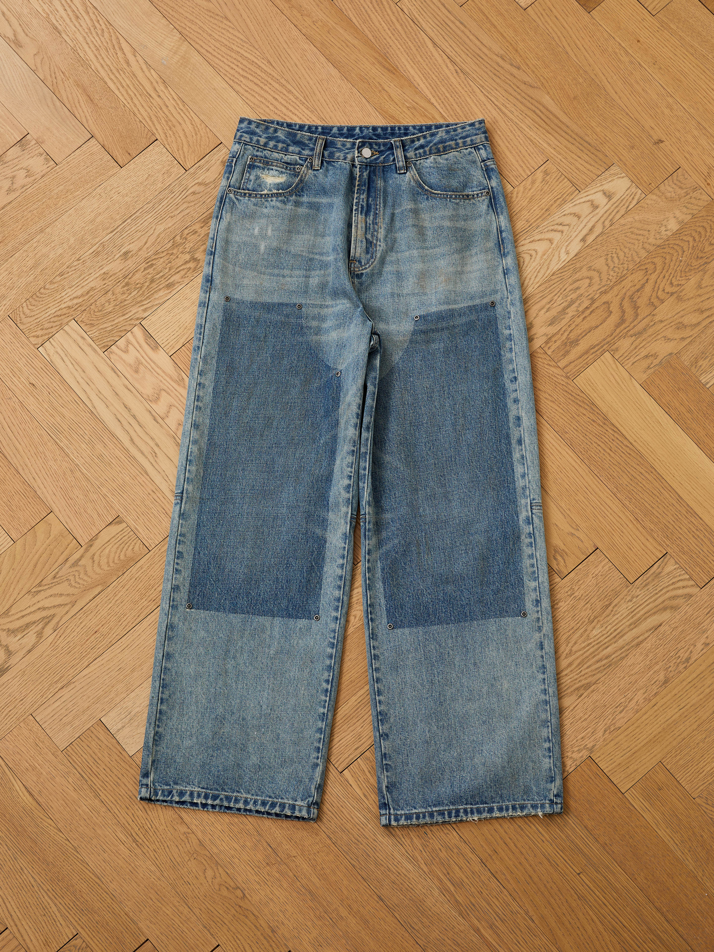 A pair of FOUND Double Knee Carpenter Jeans in blue, featuring large dark rectangular patches on the front thighs and knees, laid flat on a wooden herringbone floor.