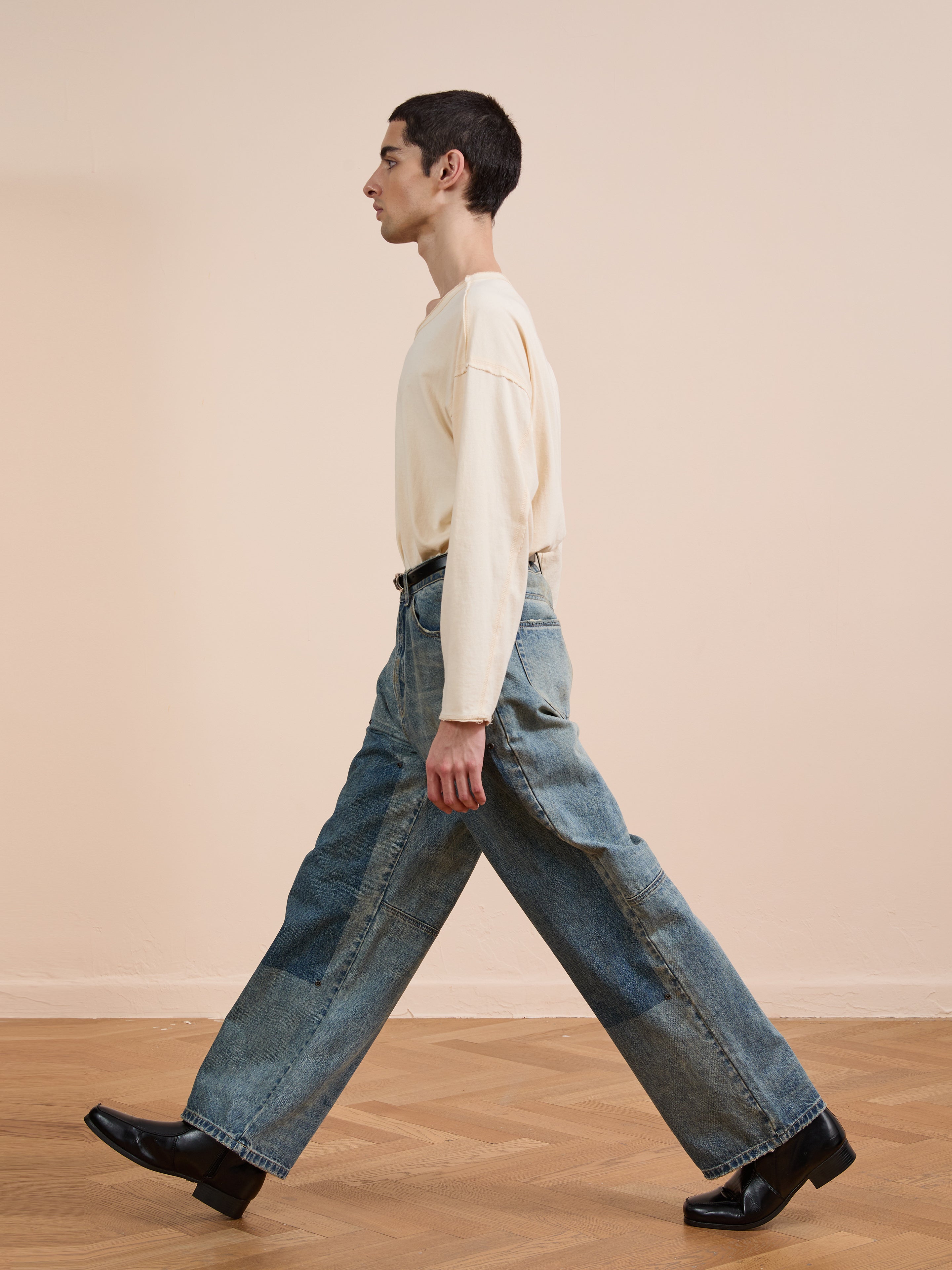 A person with short dark hair, wearing a cream long-sleeve shirt and FOUND Double Knee Carpenter Jeans, walks indoors on a wooden floor against a light beige wall.