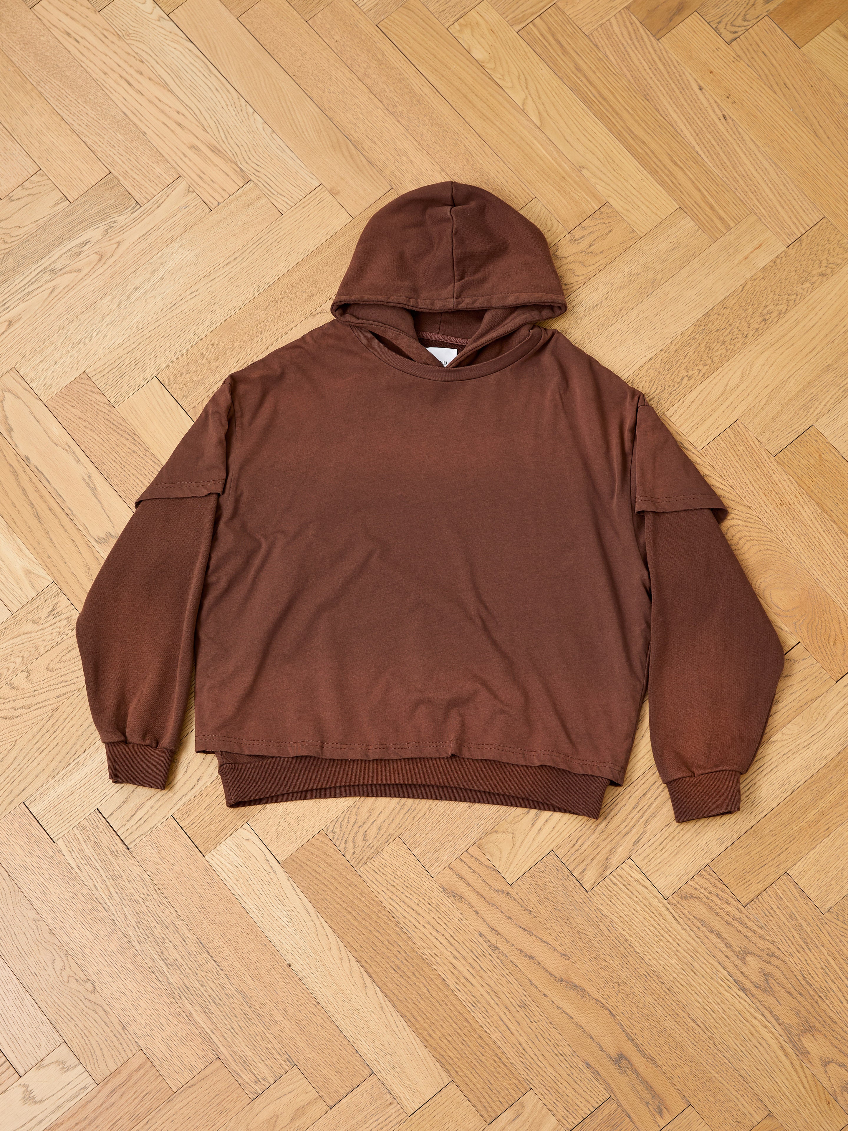 The Found Double Layer Hoodie, crafted from french terry cotton with long sleeves in brown, is displayed flat on a light wood parquet floor for a vintage effect.