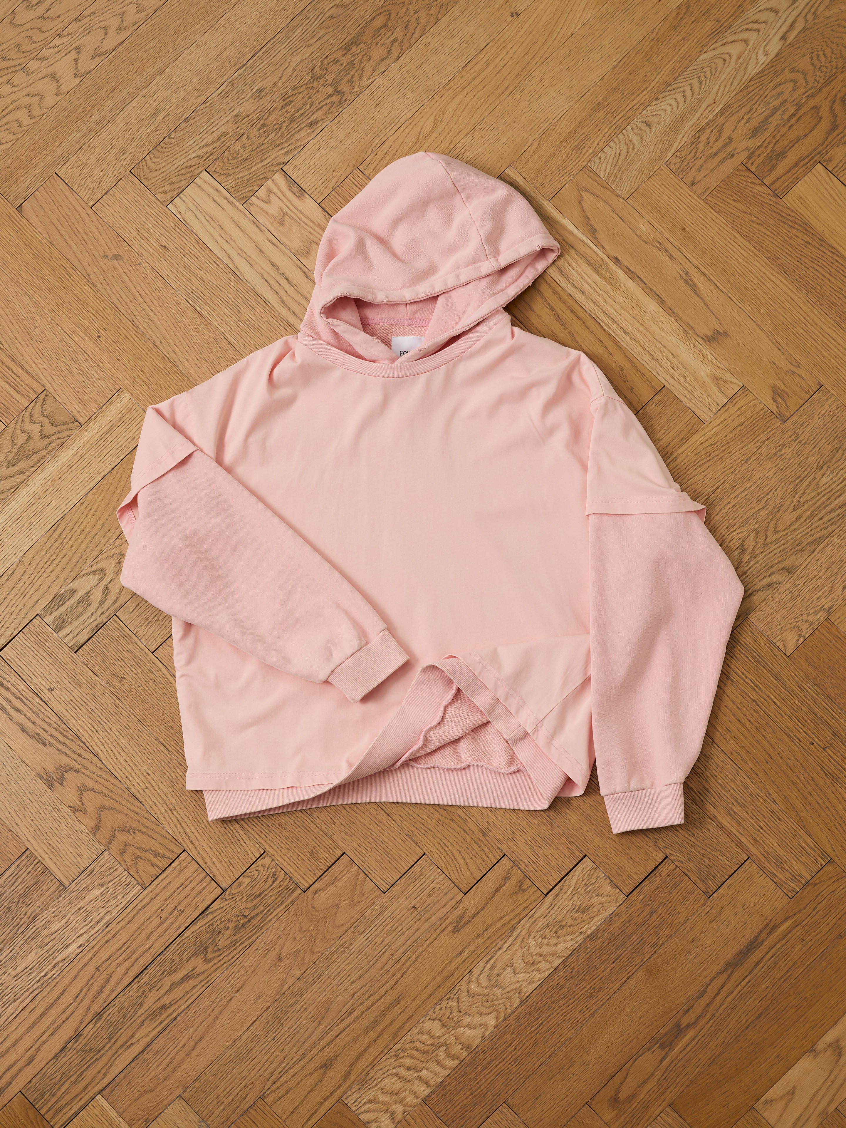 A Double Layer Hoodie by Found in light pink with a unisex relaxed fit is laid flat on a wooden herringbone floor.