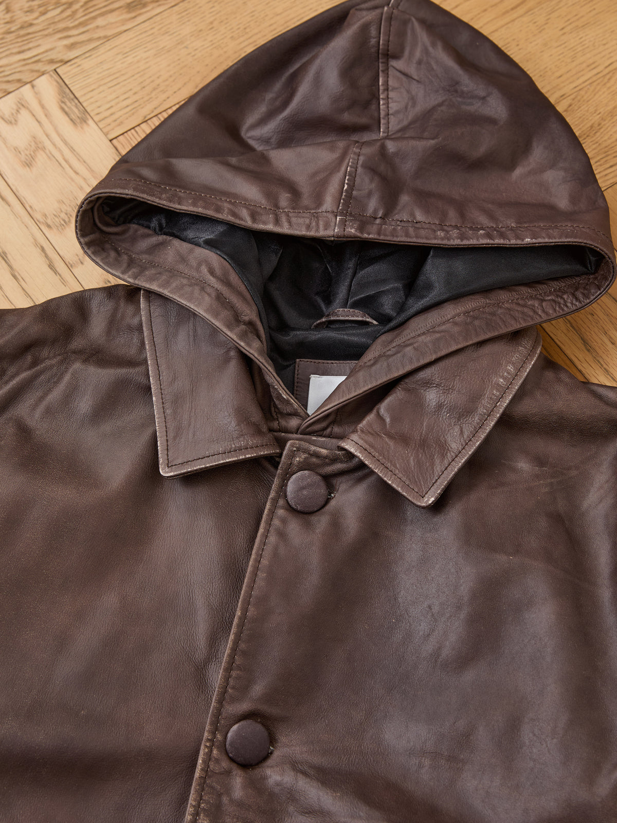 The FOUND Double Layer Leather Hooded Jacket, a unisex design with large buttons, is shown on a wooden floor. The jacket displays its interior lining and part of a clothing tag.