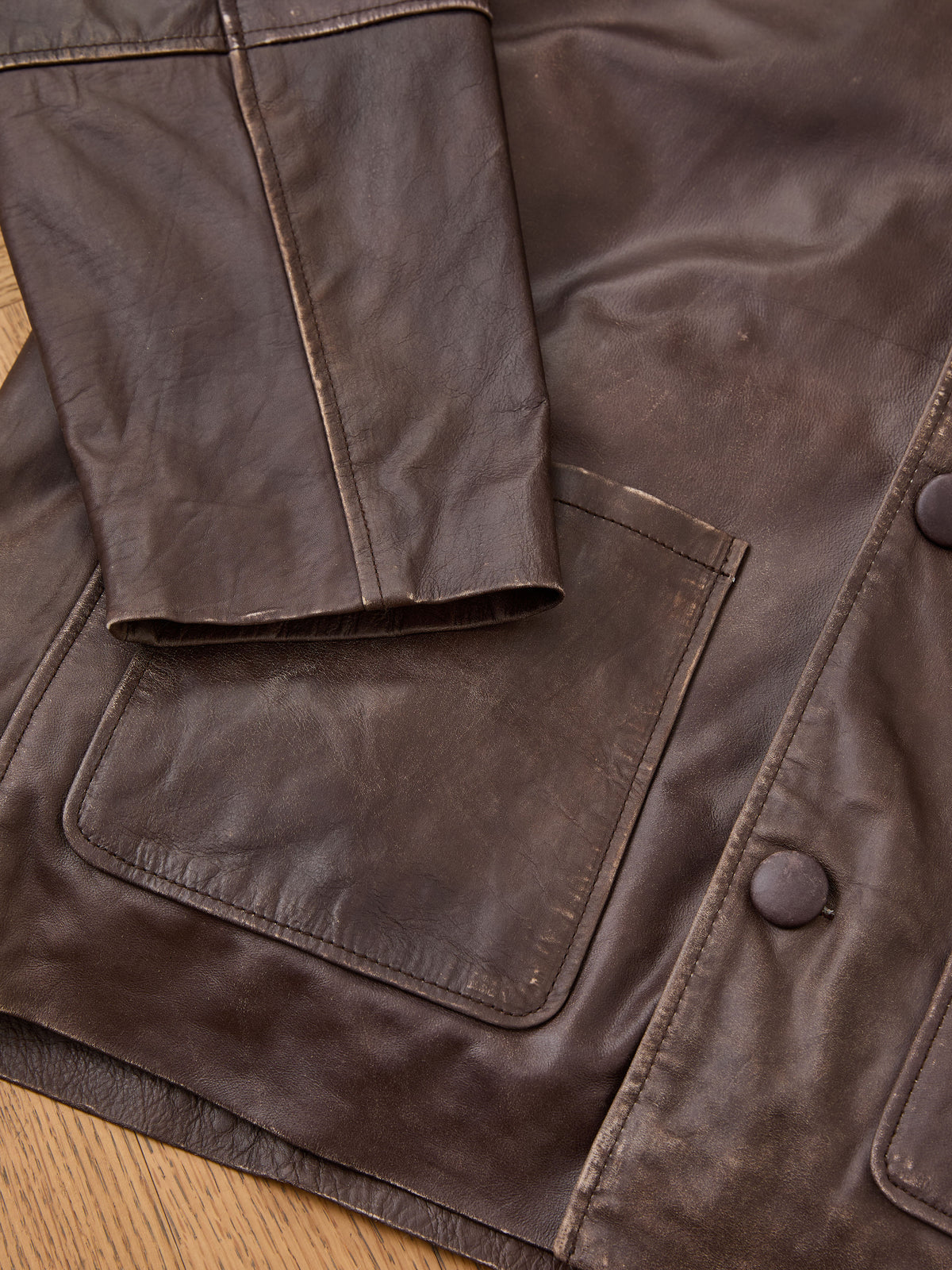 Close-up of the FOUND Double Layer Leather Hooded Jacket, highlighting a sleeve, square front pocket, and two large buttons, displayed on a wooden surface.