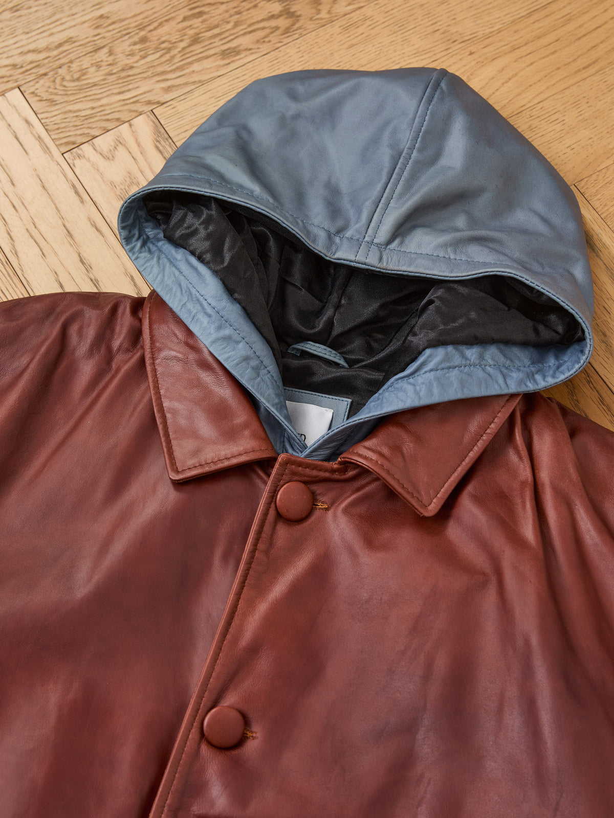 A FOUND Double Layer Leather Hooded Jacket in brown, featuring a blue hood, is laid flat on a wooden floor.
