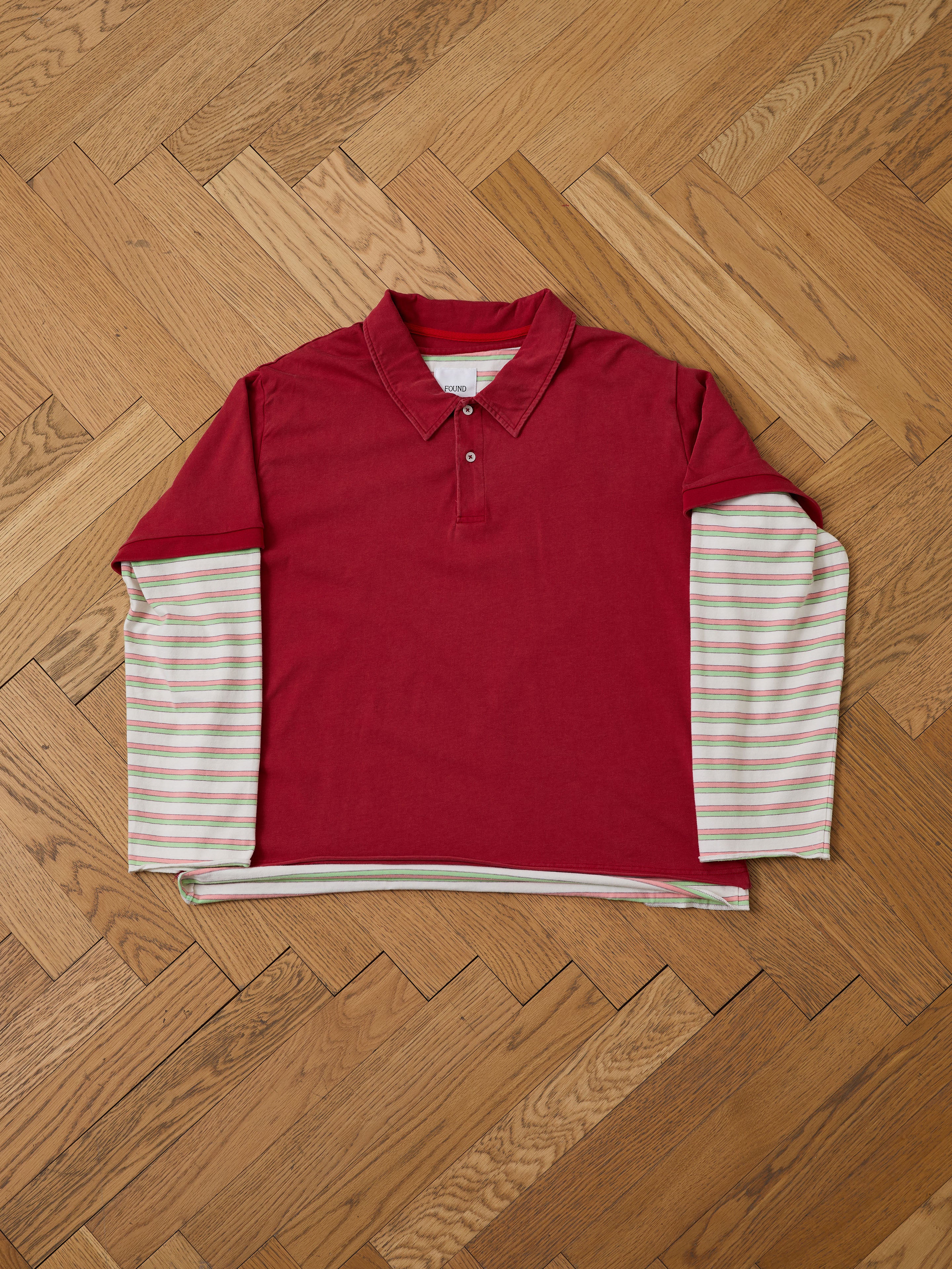 The FOUND Double Layer Stripe Polo features a unisex relaxed fit with a red collared polo over a long-sleeve white shirt with light stripes, styled in vintage athletic sportswear and displayed on a wooden herringbone floor.