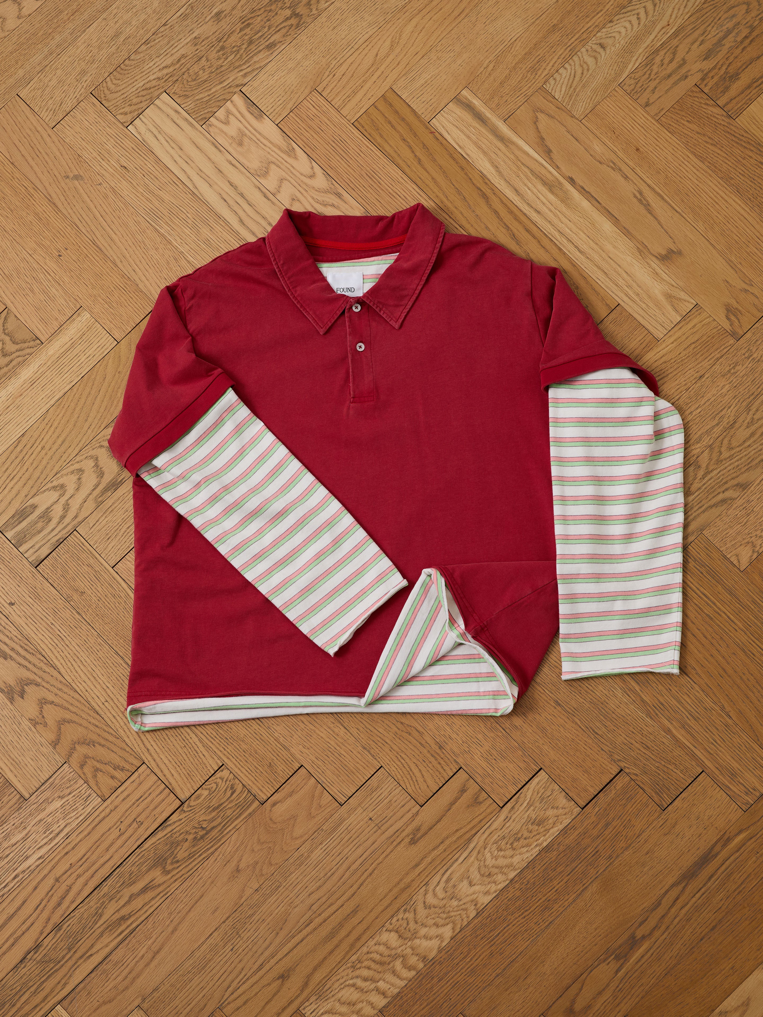 The FOUND Double Layer Stripe Polo, inspired by vintage athletic sportswear, features red fabric with white, green, and pink striped long sleeves and is laid flat on a herringbone wood floor.