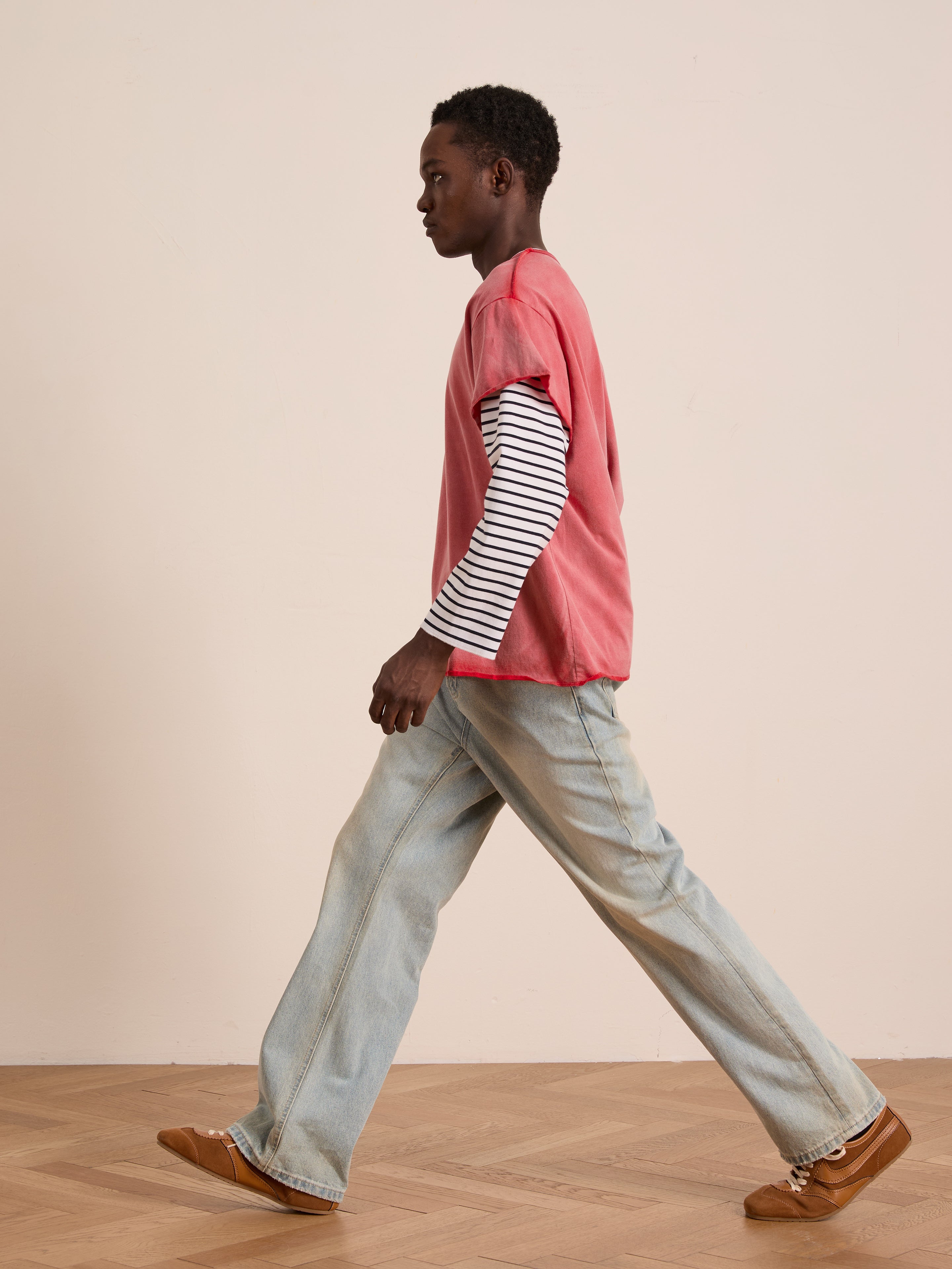 A person walks indoors in sunfade wash light blue jeans, brown shoes, and a FOUND Double Layer LS Tee—a red T-shirt over a black-and-white striped long sleeve—styled with a unisex relaxed fit.