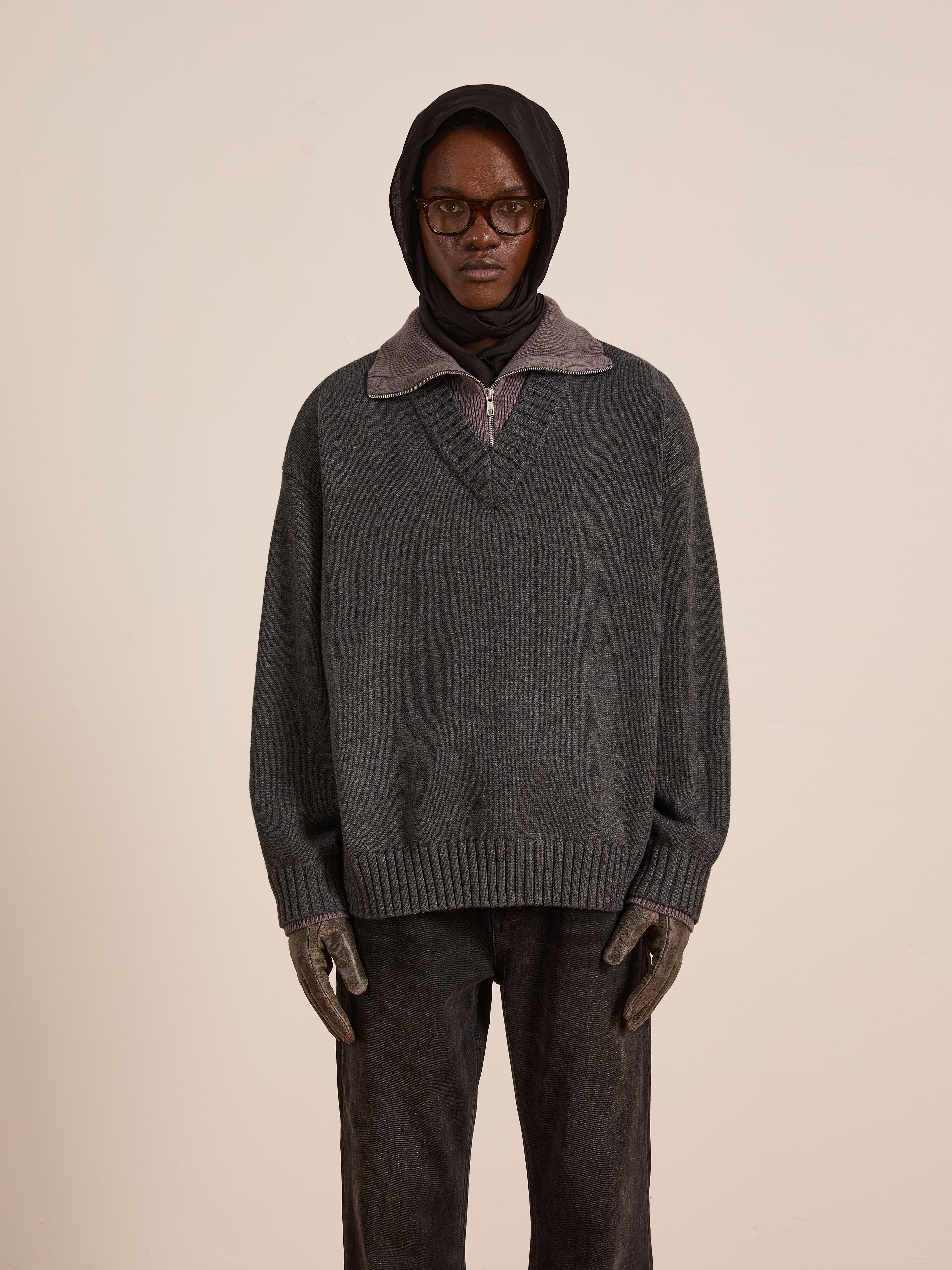 A person in FOUND’s Double Layer Sweater—an oversized gray wool blend V-neck layered over a collared shirt—paired with dark jeans, gloves, a black headscarf, and dark glasses stands against a plain light background.