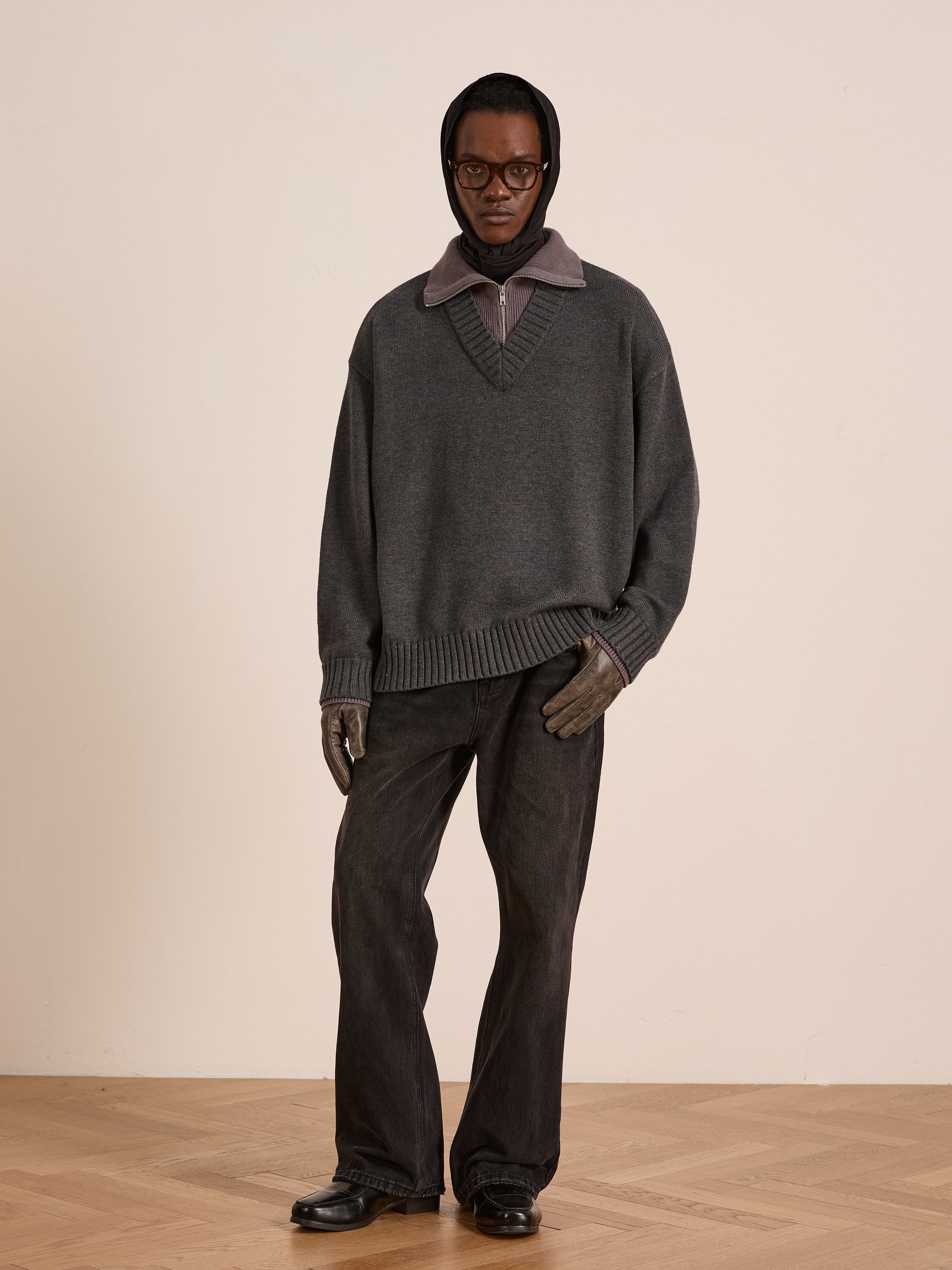 A person indoors wears dark glasses, a head covering, and the FOUND Double Layer Sweater—a gray wool blend V-neck—over a collared shirt with dark pants, gloves, and black shoes, showing ideal cold-weather layering on a wooden floor.