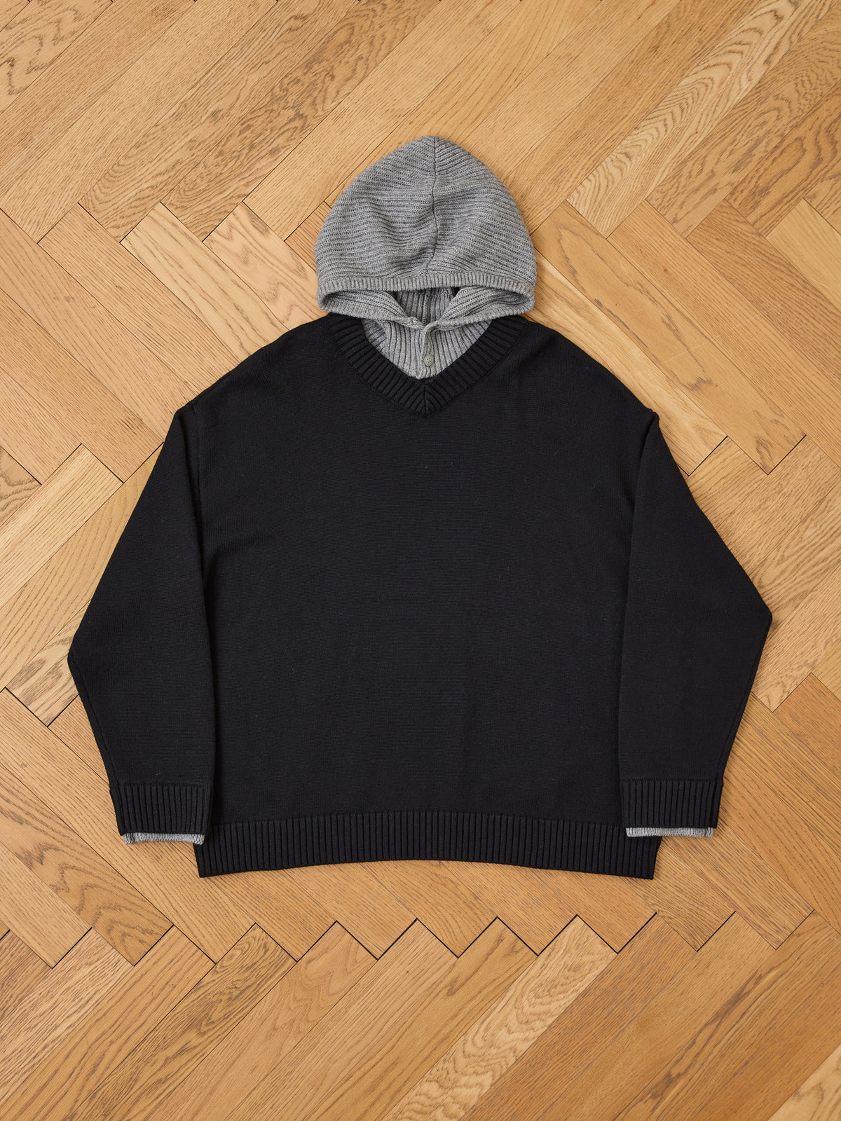 A FOUND Double Layer V Neck Knit Sweater in black, layered over a gray hooded sweatshirt, is laid flat on a light wooden herringbone floor.
