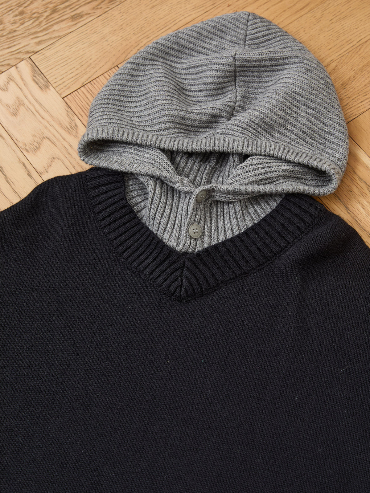 The FOUND Double Layer V Neck Knit Sweater in black features a ribbed V-neck, gray attached hood with button, shown on a wooden floor.