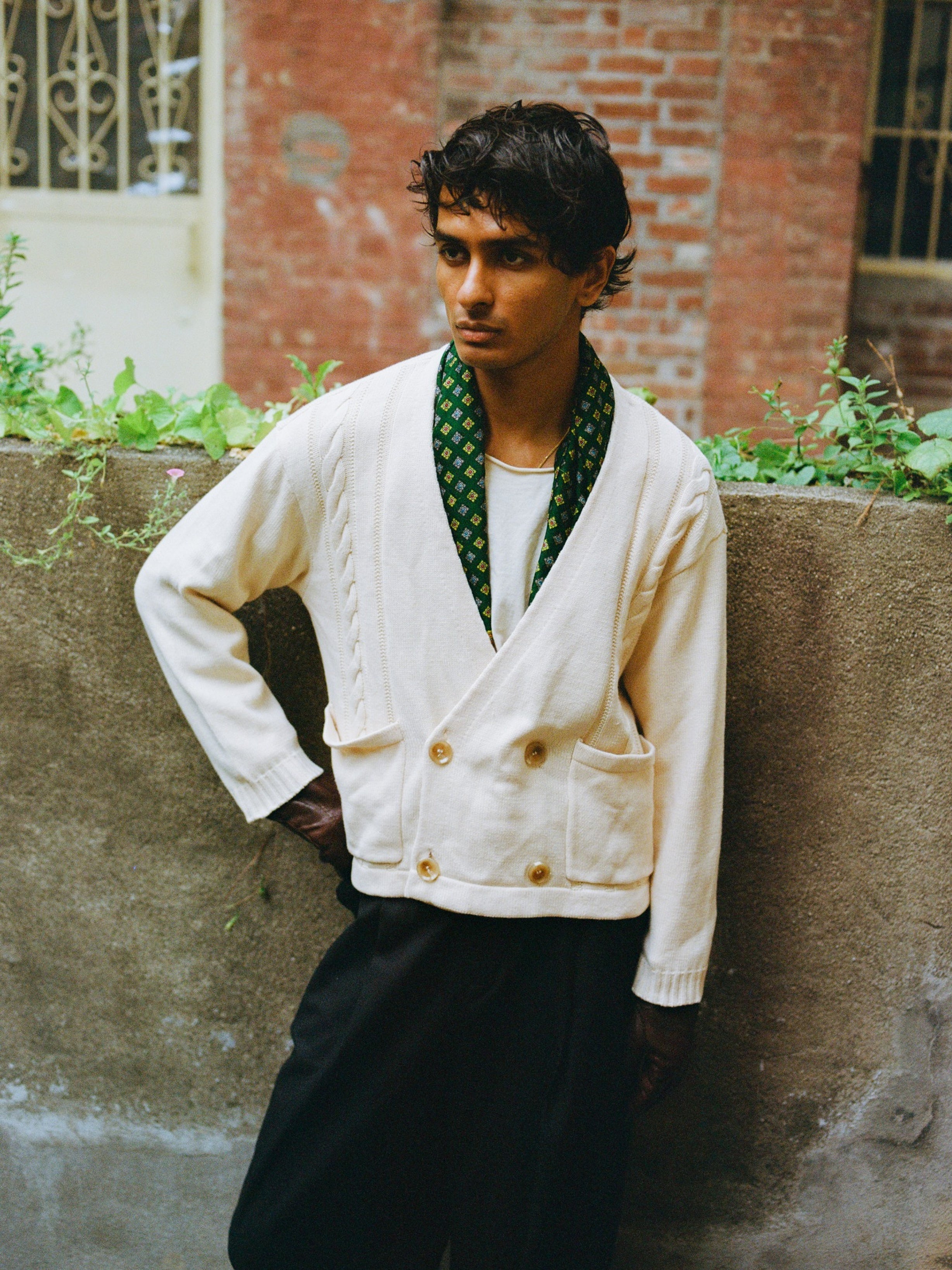 A person stands outdoors against a concrete wall, wearing the FOUND Double Breasted Knit Cardigan in white over a white shirt, black pants, and a patterned green scarf draped around their neck.