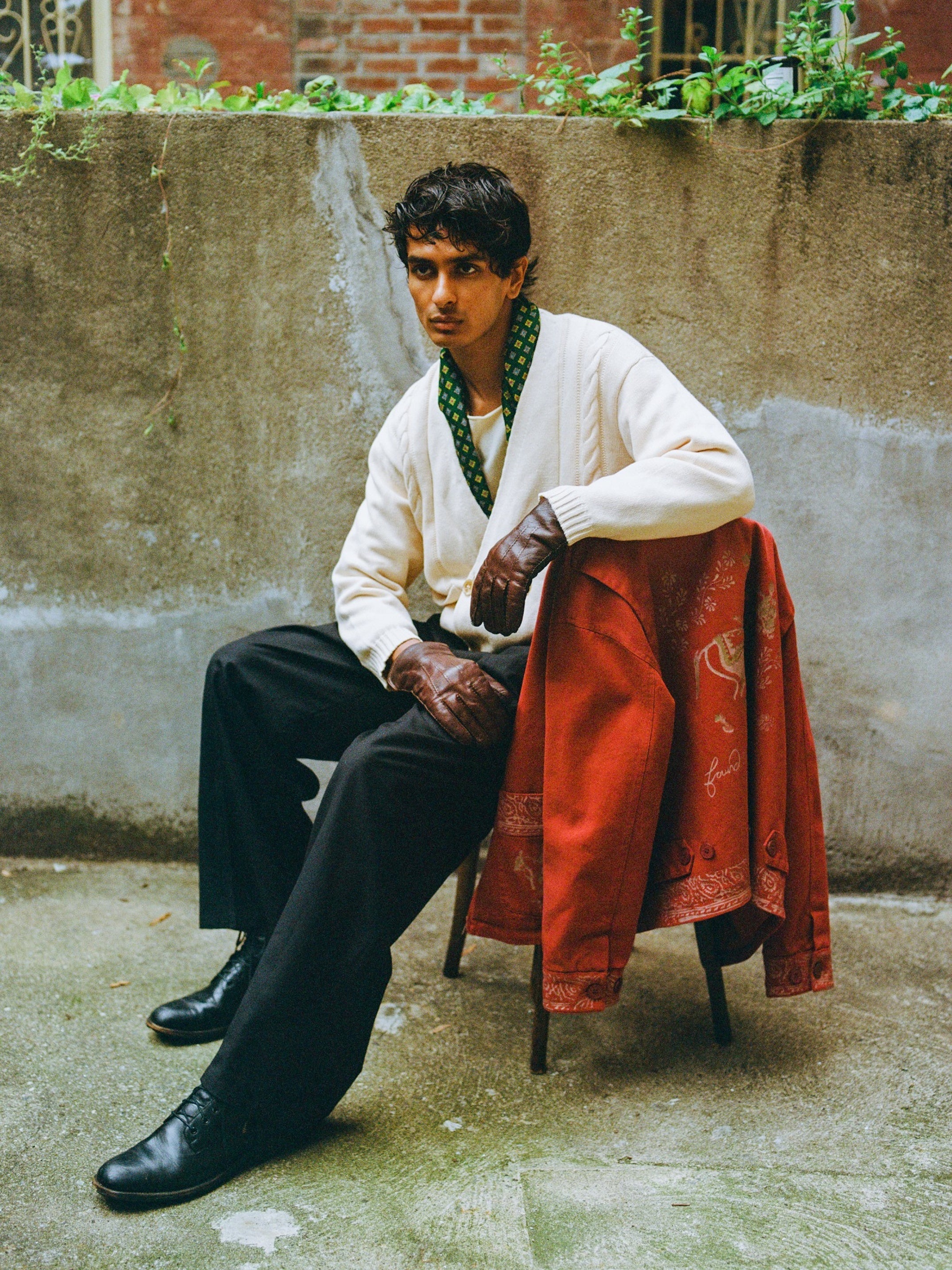 A man sits outdoors on a chair, wearing a white shirt, dark pants, gloves, and a green scarf. A FOUND Double Breasted Knit Cardigan is draped over the chair.