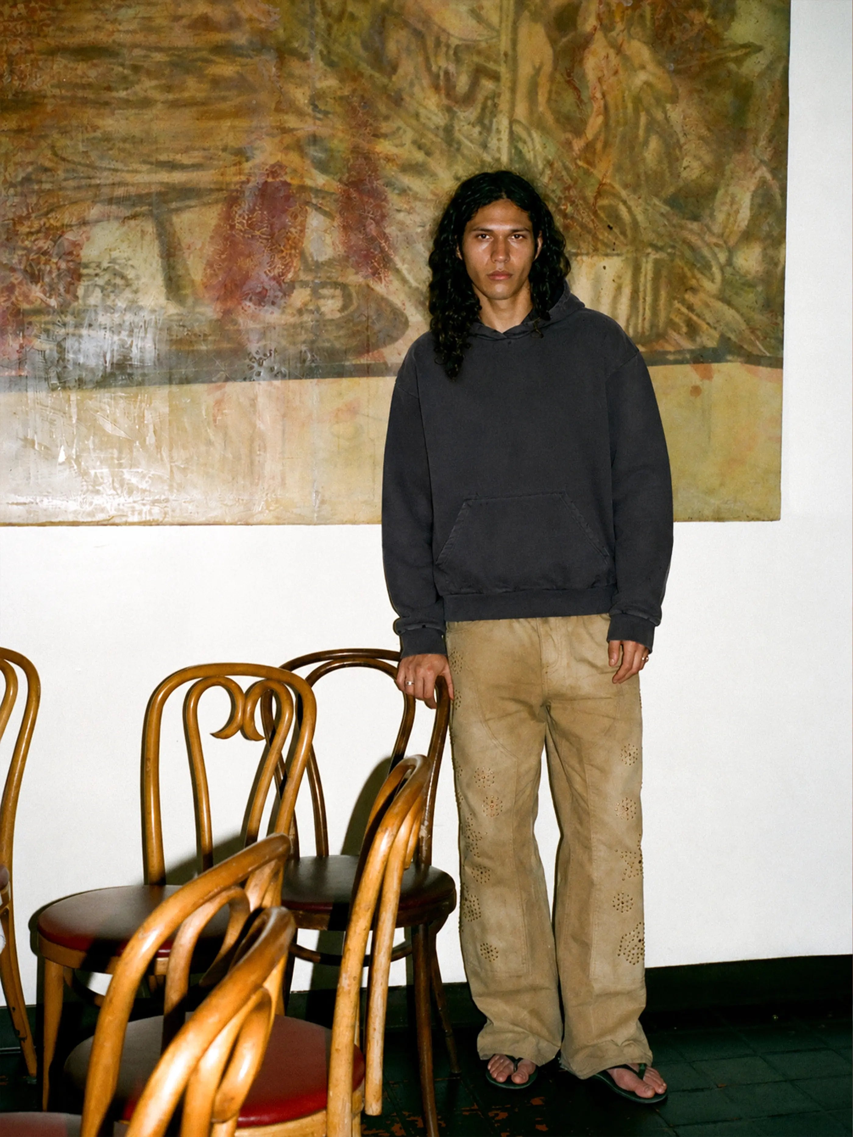 A person with long curly hair stands indoors by wooden chairs, wearing the FOUND Distressed Pullover Hoodie, tan pants, and flip-flops; a large abstract painting hangs behind them.