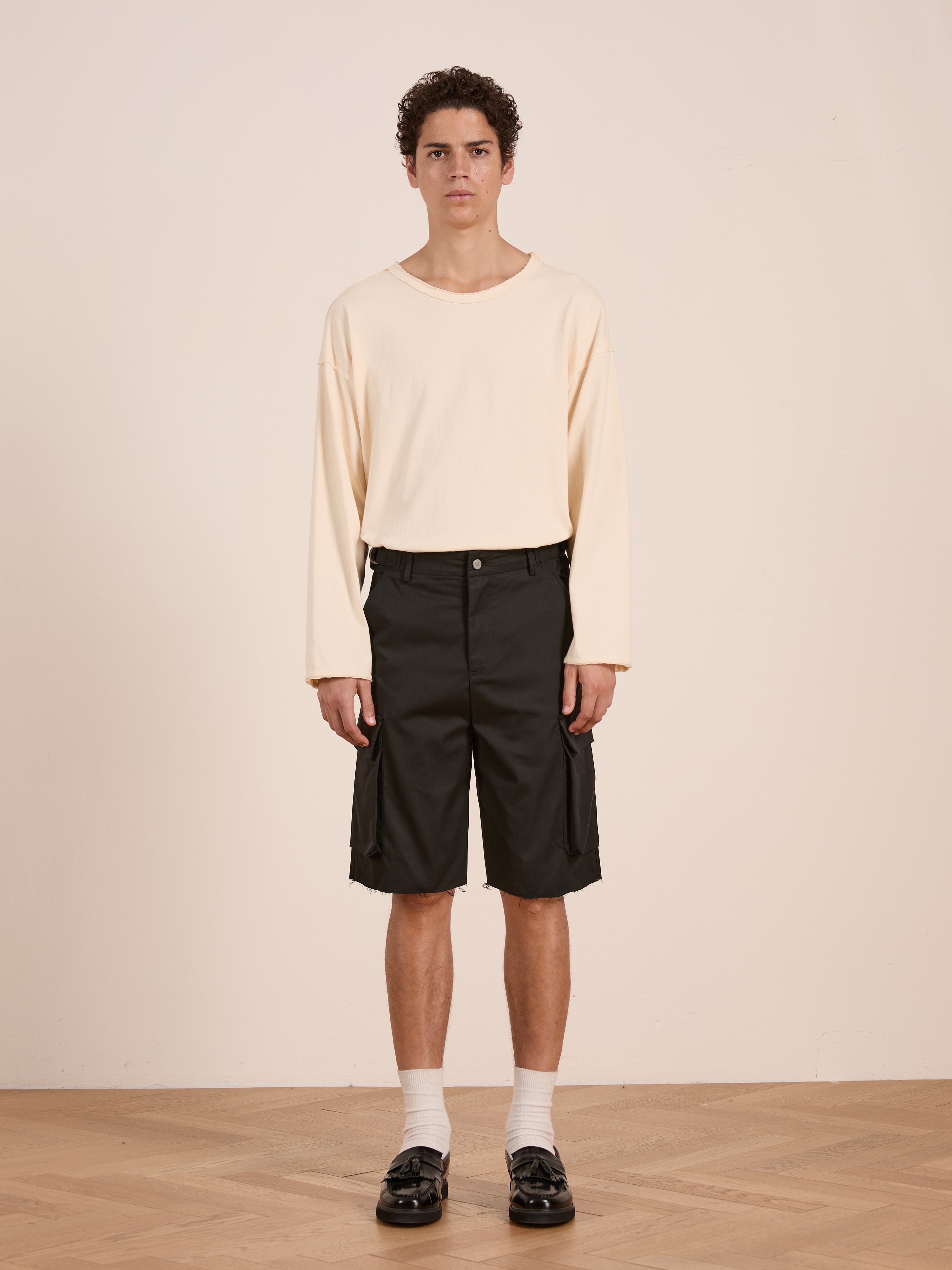 Indoors, a person wears a loose beige long-sleeve shirt and Found Elbas Cargo Shorts with dual cargo pockets and adjustable waist tabs, paired with white socks and black loafers against a plain light wall.