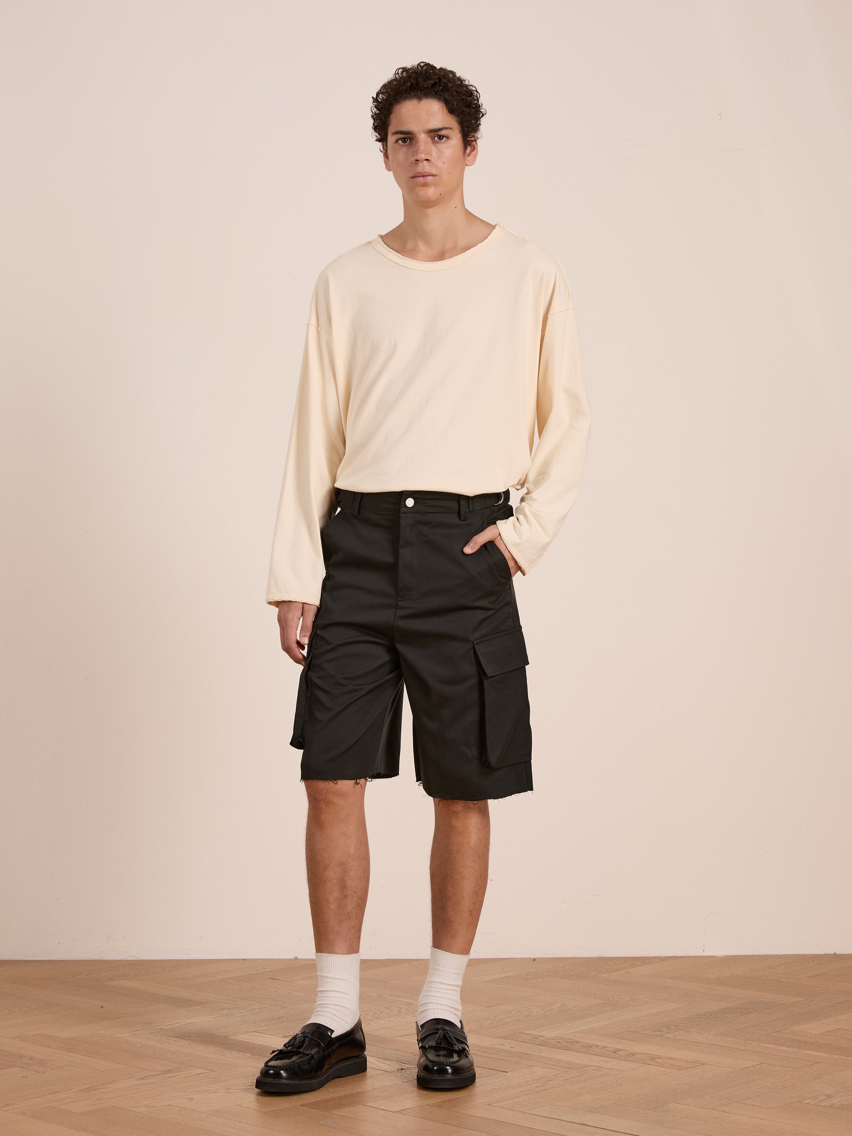 Indoors, a person wears a cream long-sleeve shirt, Found Elbas Cargo Shorts with dual cargo pockets and adjustable waist tabs, white socks, and black loafers, posing with one hand in their pocket.