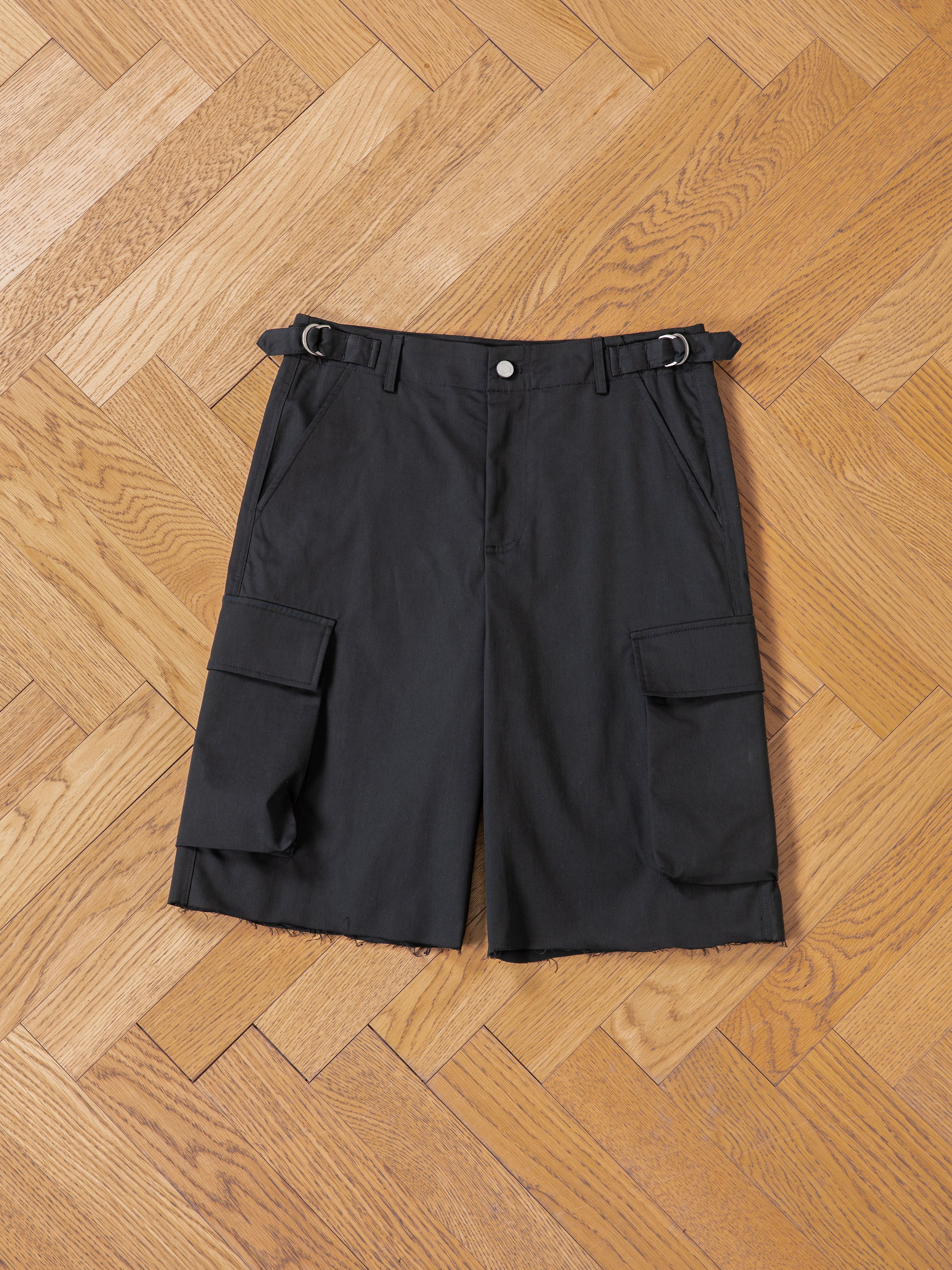 Found Elbas Cargo Shorts in black, laid flat on a light wooden herringbone floor, featuring dual cargo pockets and adjustable waist tabs.