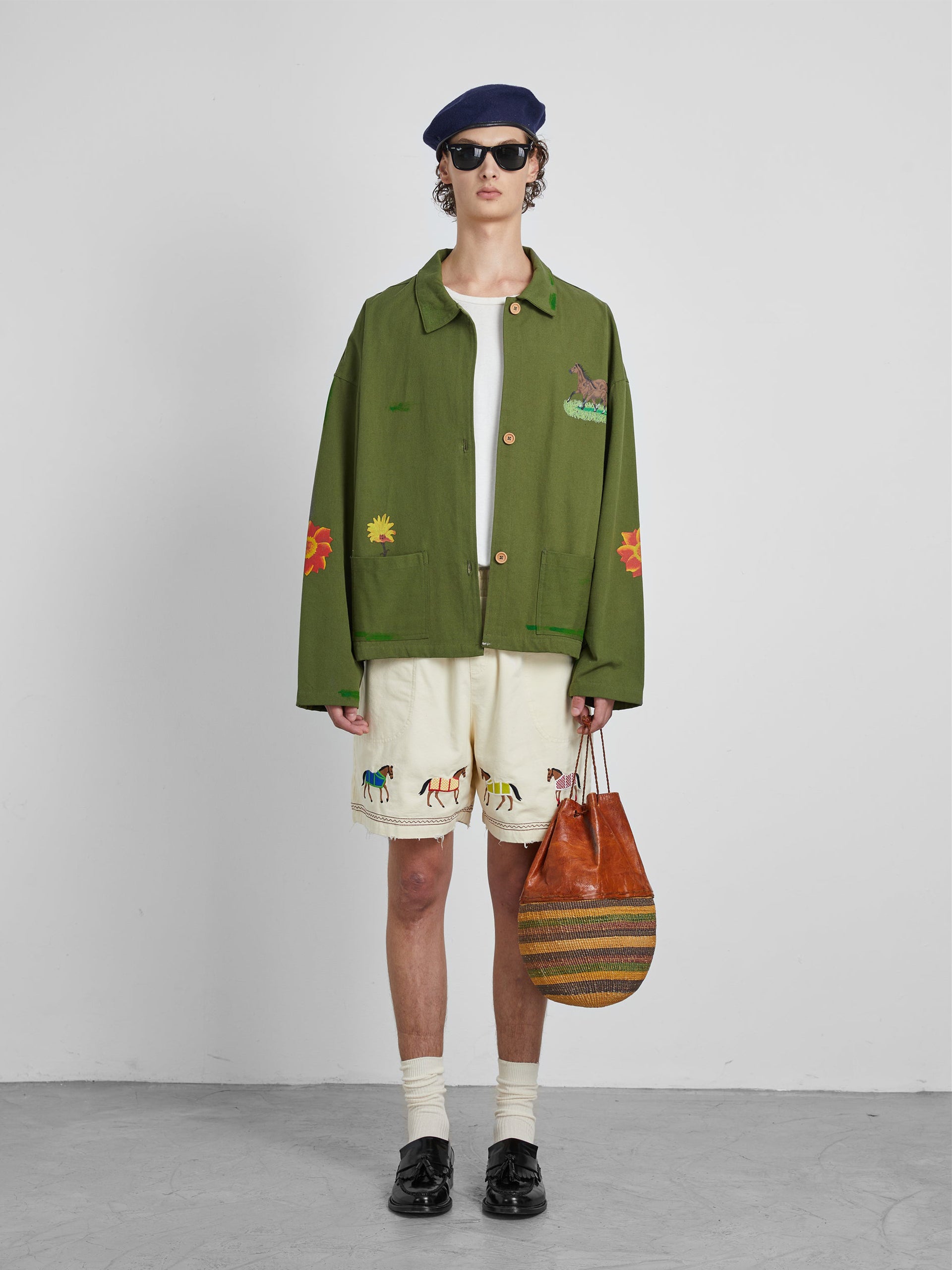 A person wearing a green jacket, the Profound Embroidered Horse Shorts in beige cotton twill, black shoes, and a beret holds a striped brown bag.