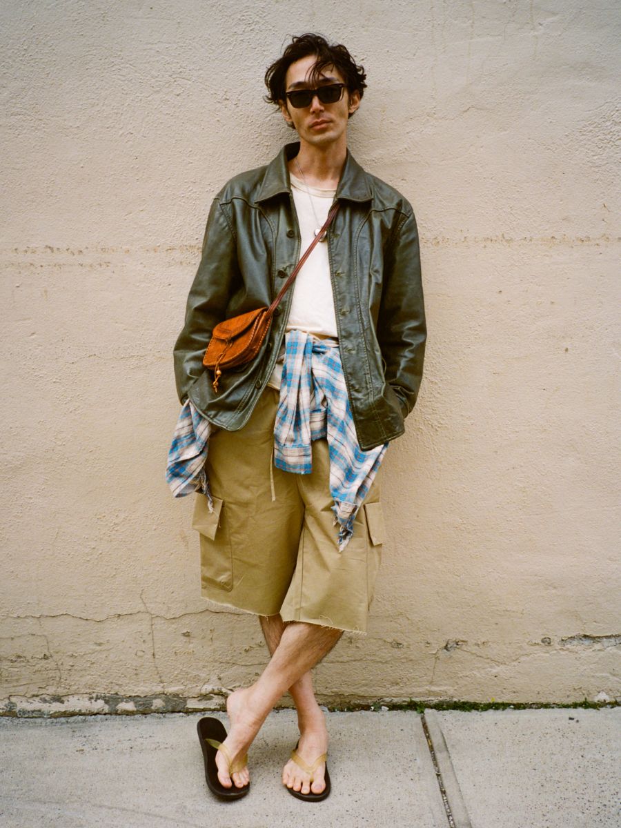 A man wearing sunglasses stands against a beige wall in a FOUND Leather Field Jacket, tan cargo shorts, a plaid shirt tied at his waist, flip-flops, and a brown crossbody bag.