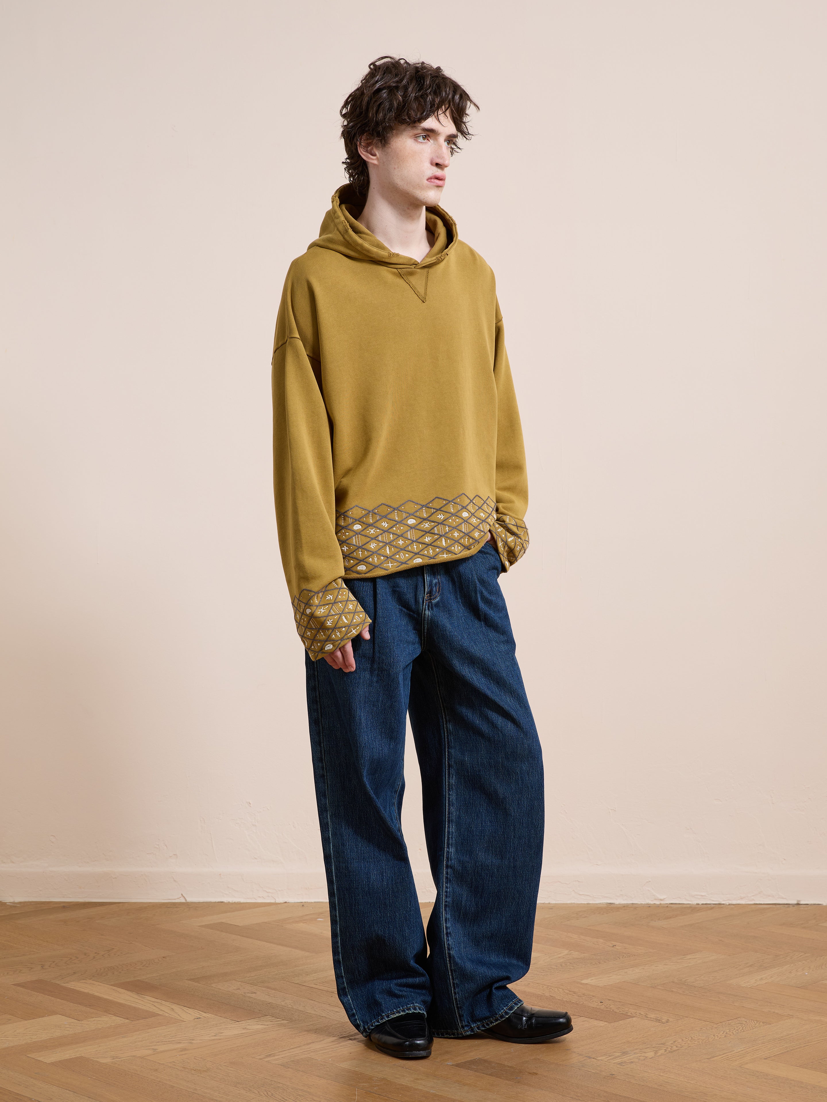 A person stands on a wooden floor against a plain beige wall, wearing the FOUND Embroidered Border Hoodie in mustard with patterned cuffs and hem, paired with wide-leg blue jeans and black shoes.
