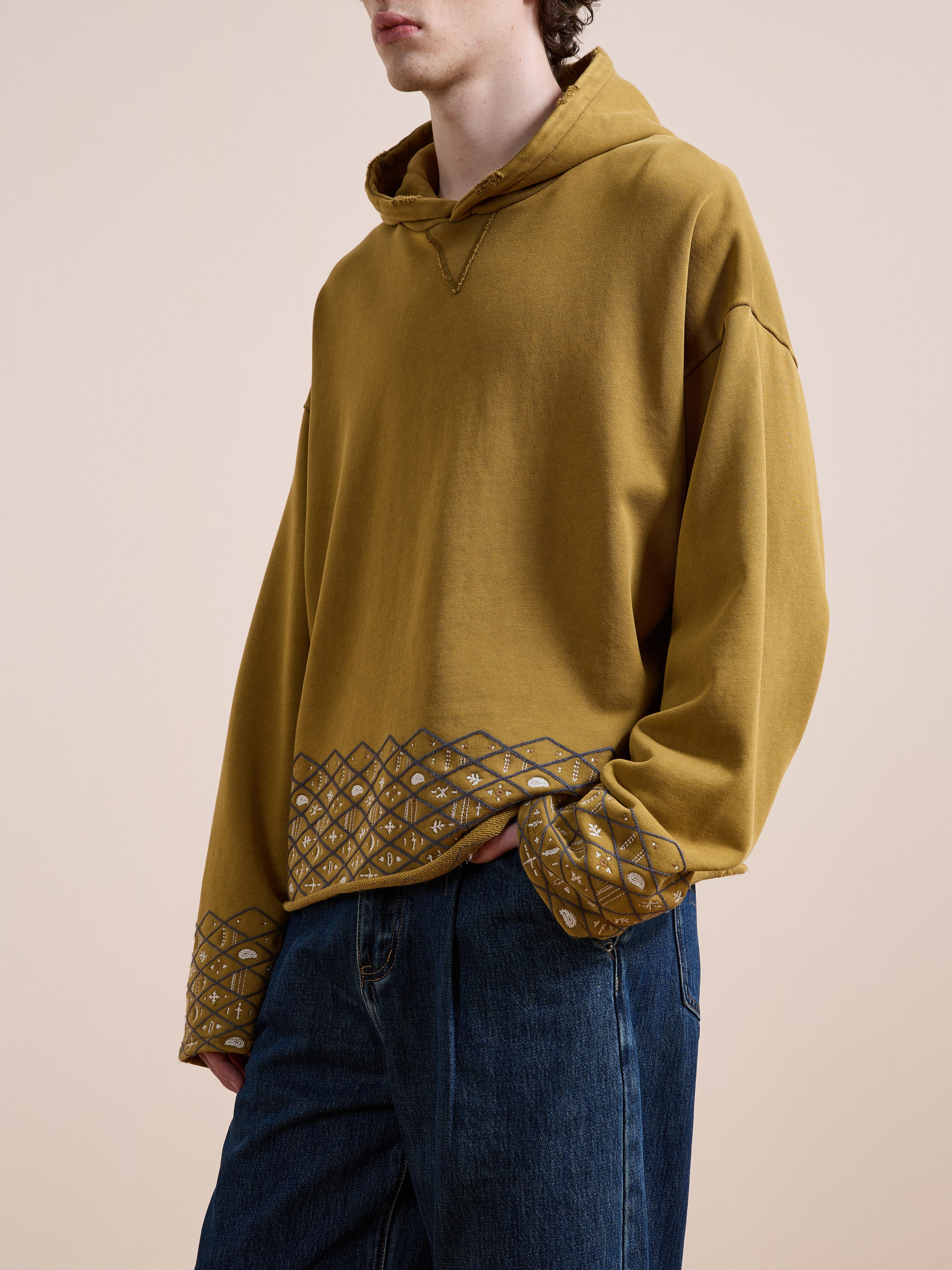A person wears the FOUND Embroidered Border Hoodie, a vintage-style cotton hoodie in mustard yellow featuring geometric embroidered hems and cuffs, paired with blue jeans against a neutral background.