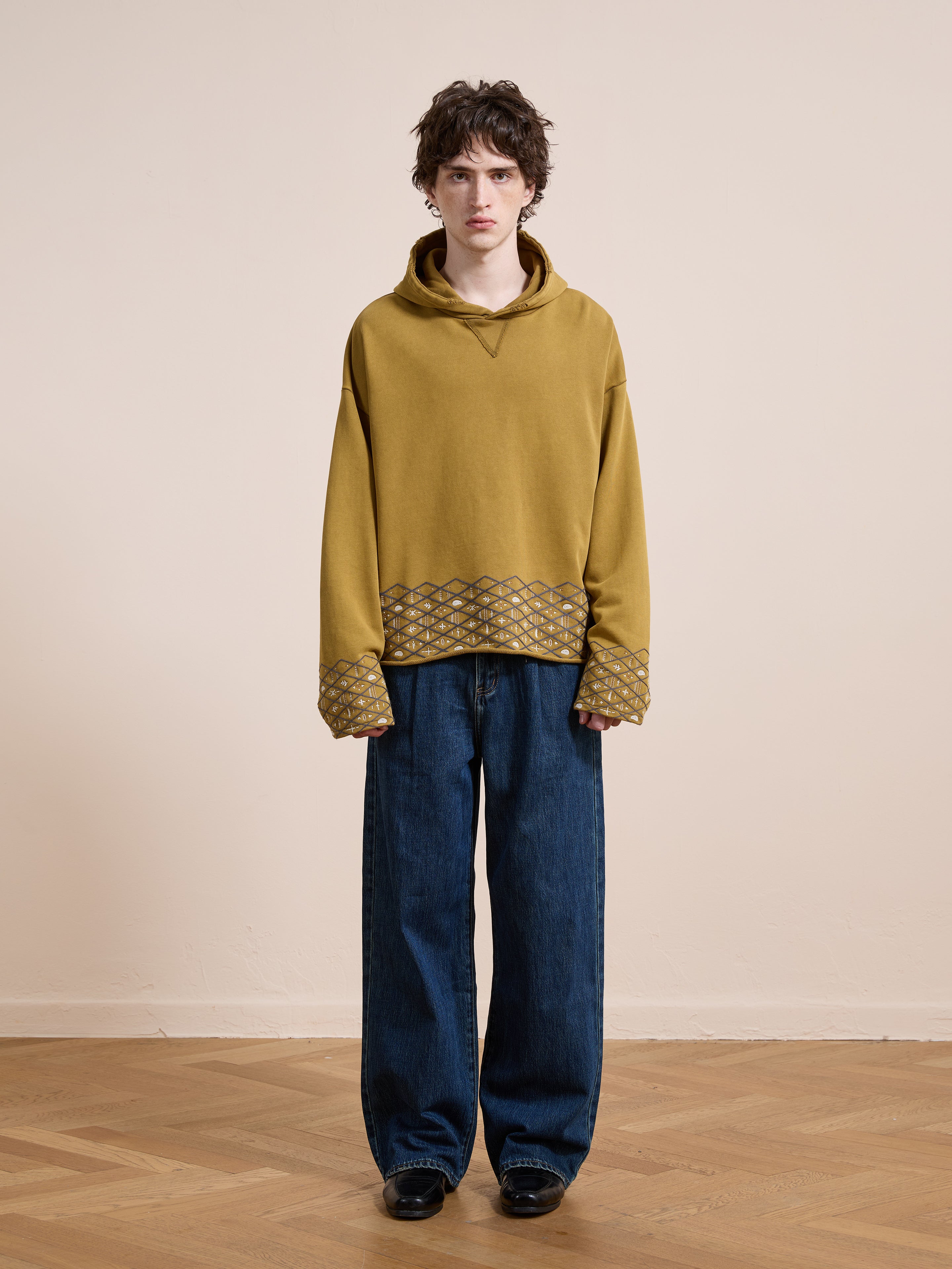 A person stands indoors against a plain wall, wearing FOUND’s Embroidered Border Hoodie in mustard yellow with patterned cuffs and hem, styled with wide-leg blue jeans and black shoes.