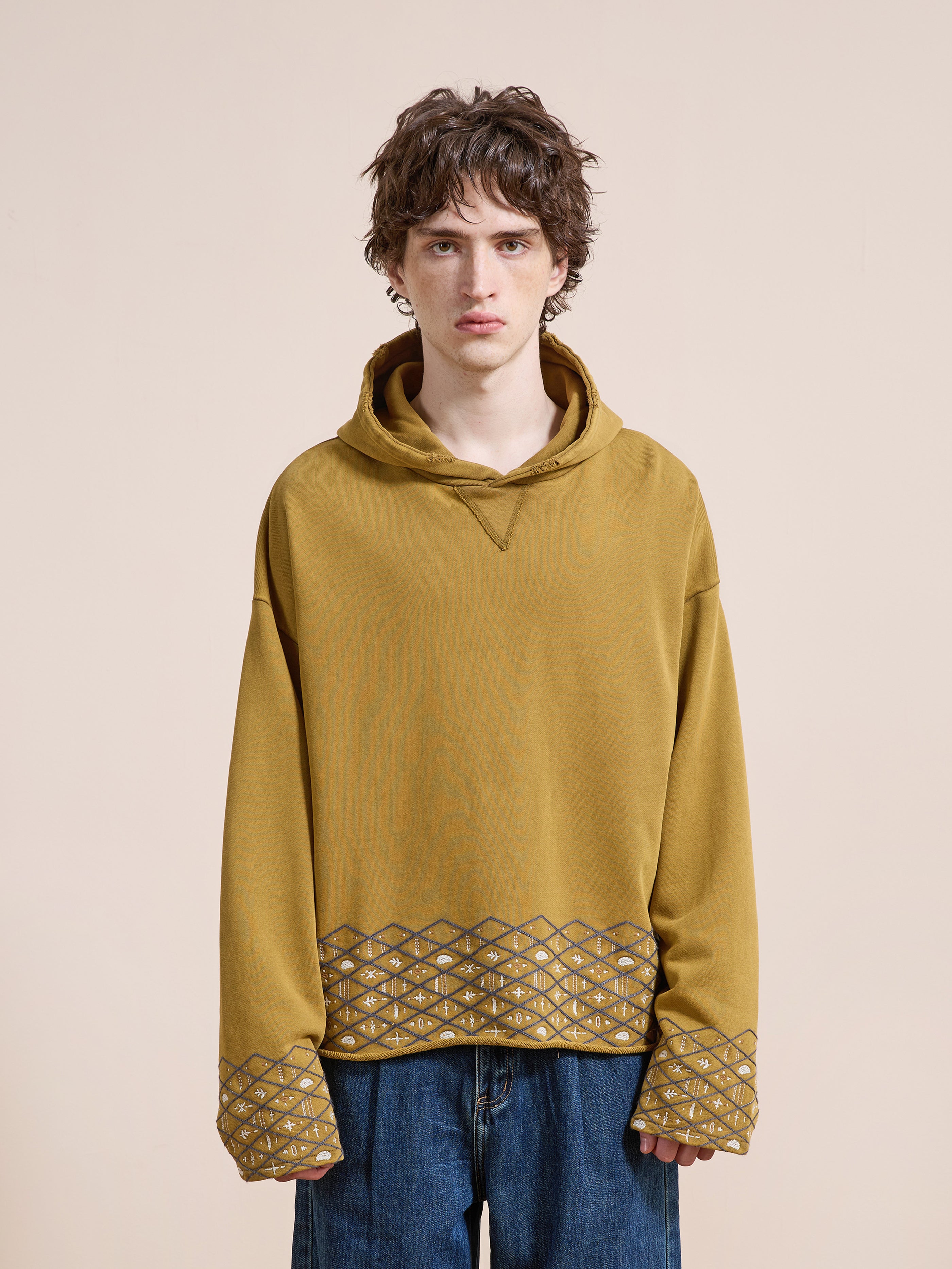 A young person with short curly hair wears the FOUND Embroidered Border Hoodie in mustard yellow, featuring geometric-patterned cuffs and hem, paired with blue jeans against a plain beige background.